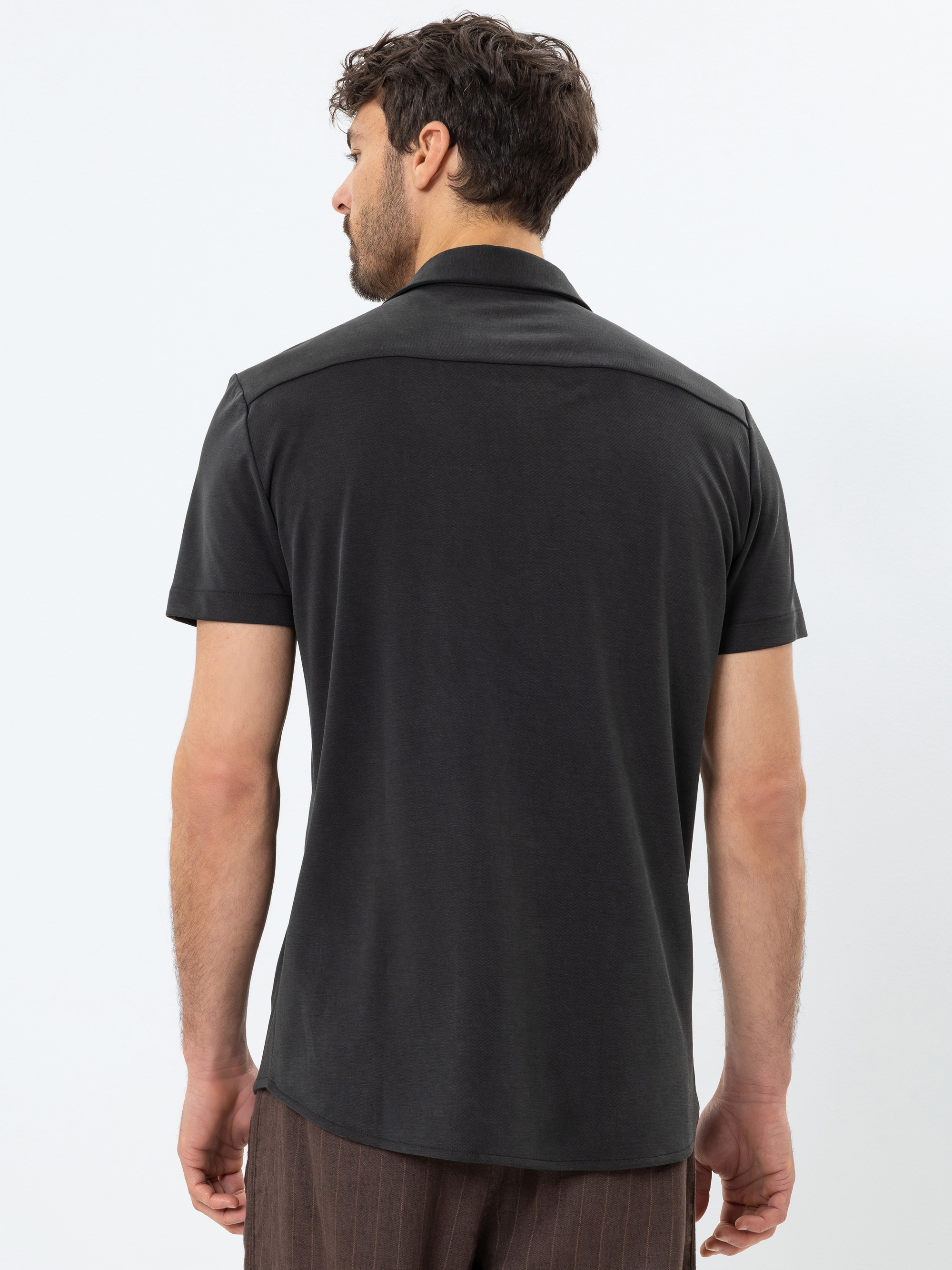 Ein Mann mit kurzen, dunklen Haaren steht mit dem Rücken zur Kamera, trägt ein kurzärmeliges, dunkles Polohemd und eine braune Nadelstreifenhose vor einem schlichten weißen Hintergrund.