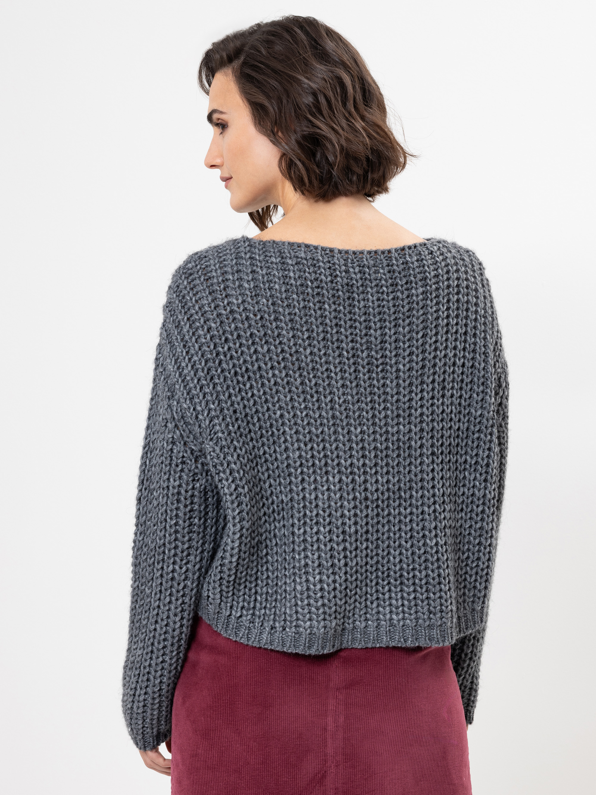 Eine Frau mit kurzen braunen Haaren steht mit dem Gesicht zur Seite. Sie trägt einen dicken, dunkelgrauen Strickpullover und einen dunkelroten Rock vor einem schlichten weißen Hintergrund.