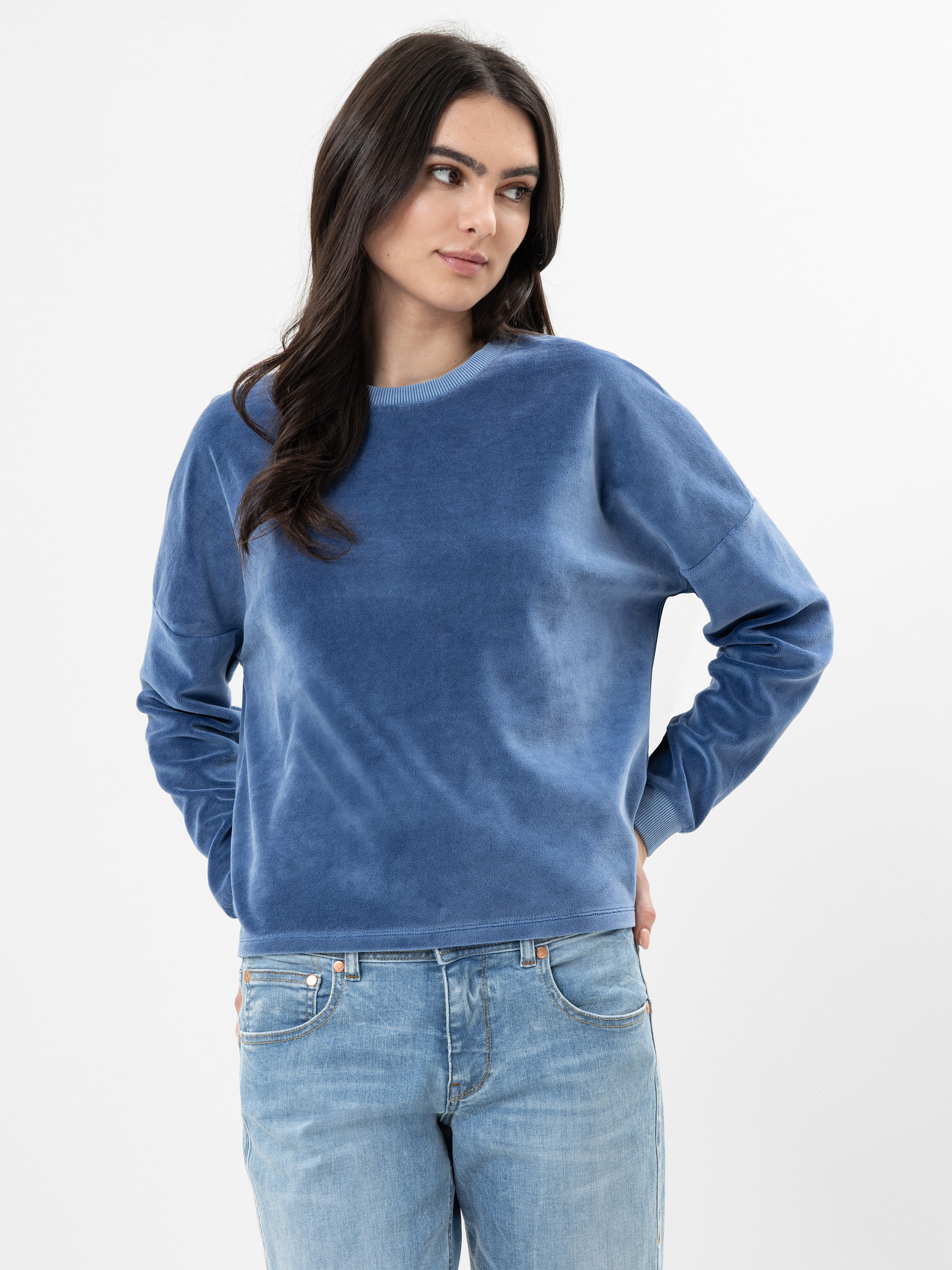 Eine Frau mit langen dunklen Haaren, die einen blauen Samtpullover und hellblaue Jeans trägt, steht vor einem schlichten weißen Hintergrund und blickt mit neutralem Blick leicht zur Seite.