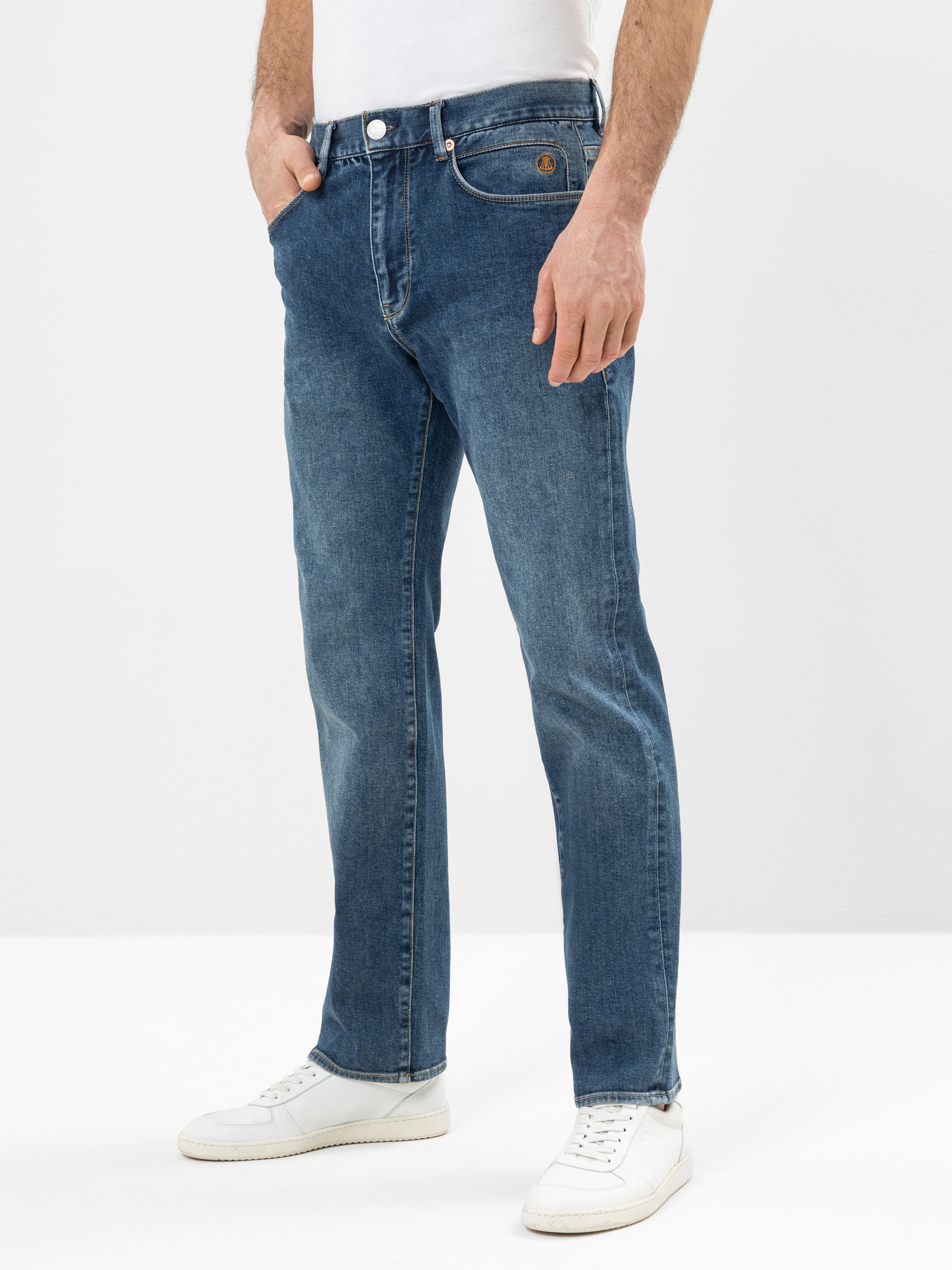 Ein Mann trägt eine blaue Jeans mit geradem Bein und den Händen in den Taschen, dazu weiße Turnschuhe und ein schlichtes weißes Hemd. Der Hintergrund ist schlicht und weiß.