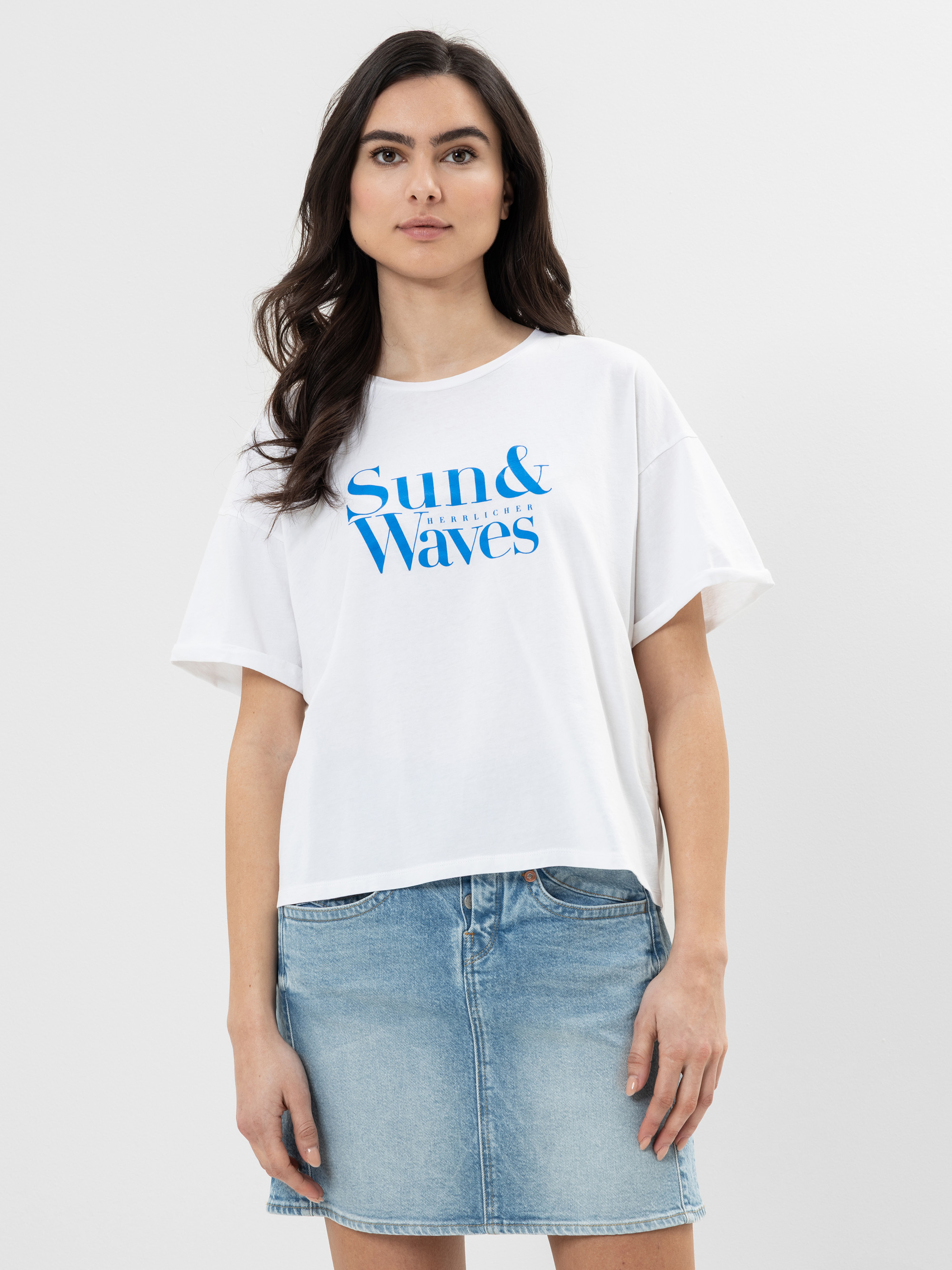 Eine Frau mit langen dunklen Haaren trägt ein weißes T-Shirt mit der blauen Aufschrift "Sun & Waves" und einen hellblauen Jeansrock. Sie steht vor einem schlichten hellgrauen Hintergrund und blickt mit neutralem Gesichtsausdruck nach vorne.