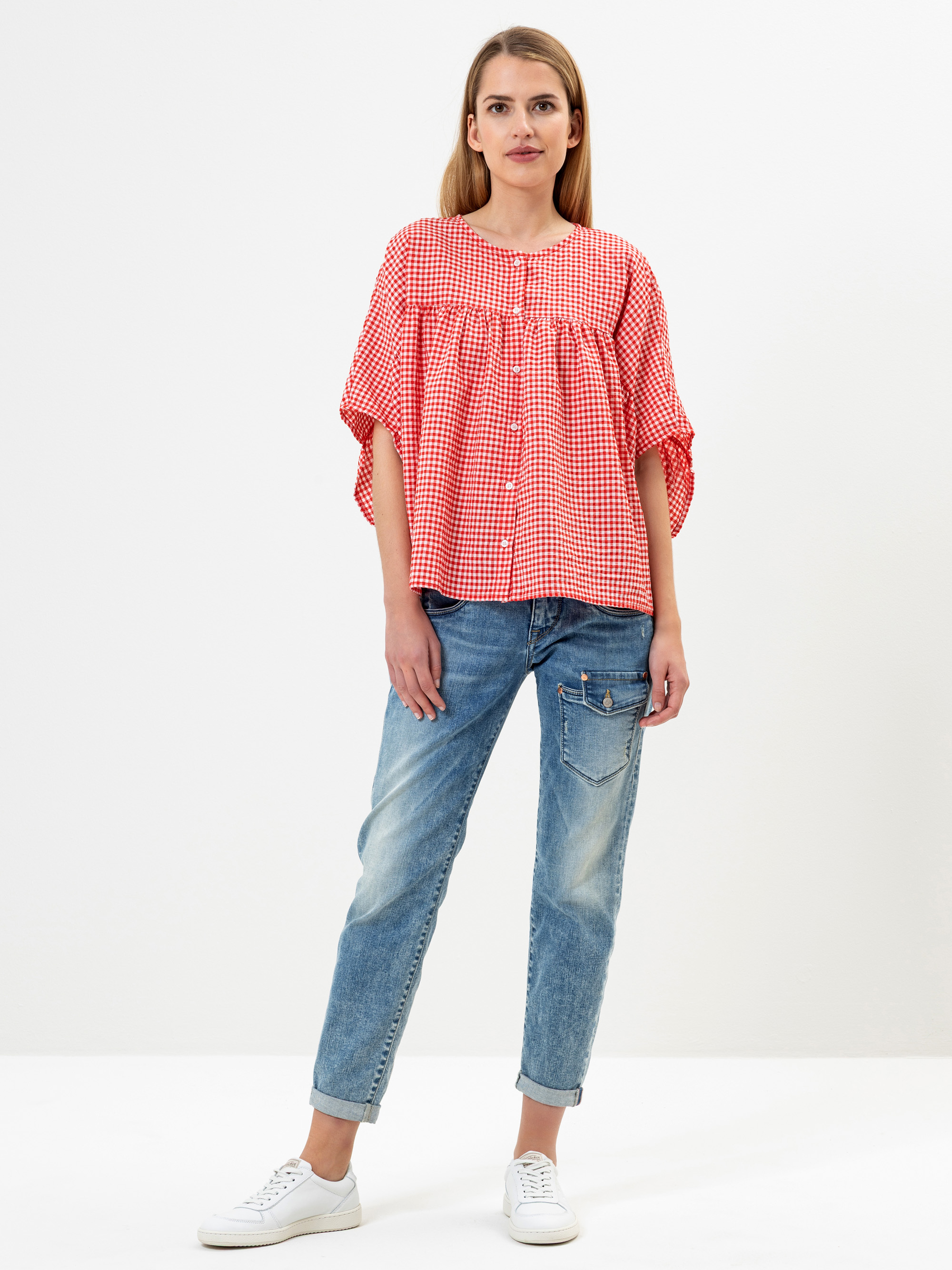 Eine Frau steht vor einem schlichten Hintergrund und trägt eine rot-weiß-karierte, lockere Bluse, blaue Jeans mit Bündchen und weiße Turnschuhe. Sie hat glattes, hellbraunes Haar und einen neutralen Gesichtsausdruck.