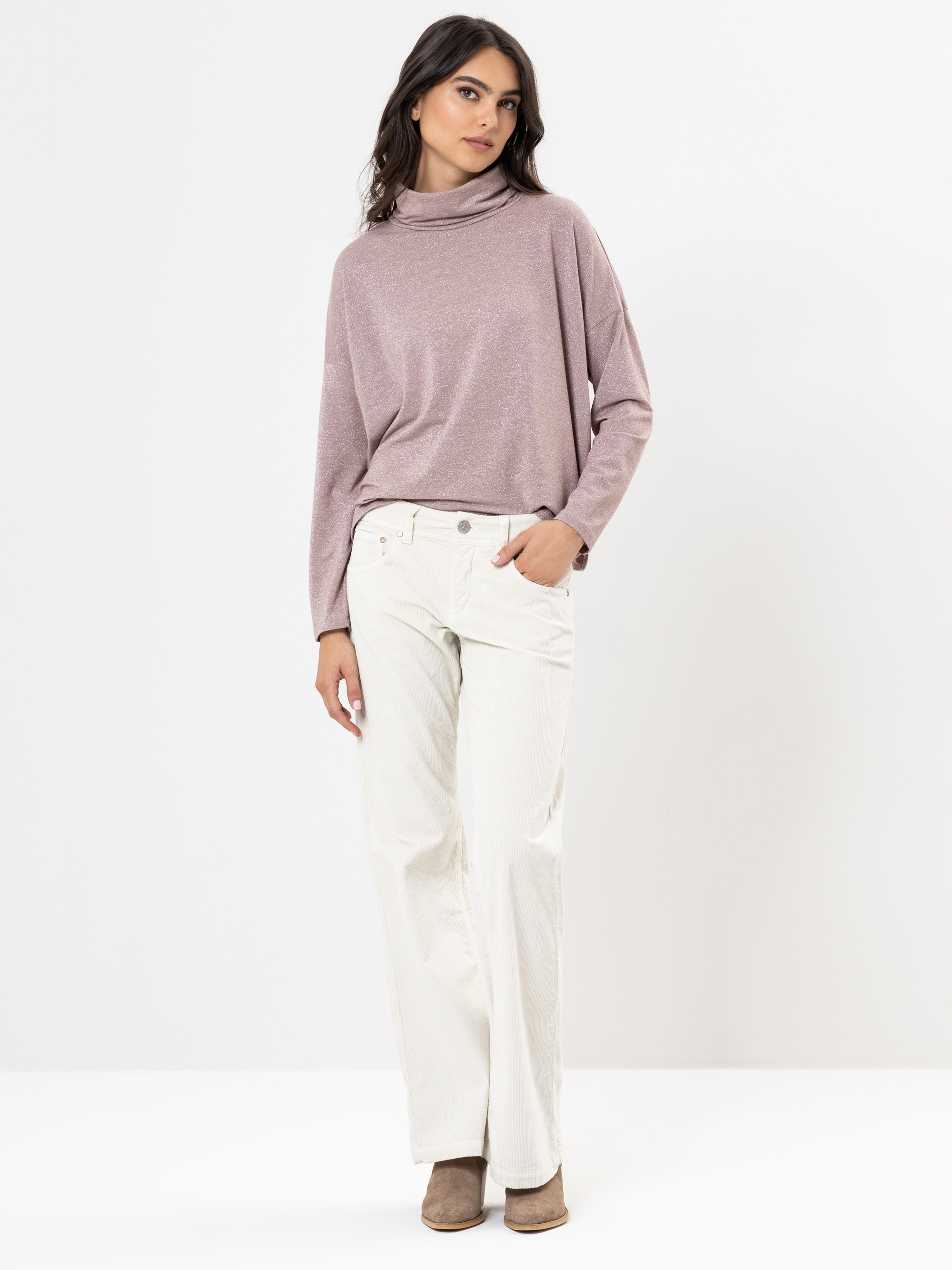 Eine Frau steht vor einem schlichten weißen Hintergrund, trägt einen hellvioletten Rollkragenpullover, eine weiße Hose mit weitem Bein und braune Stiefeletten, eine Hand in der Tasche und einen entspannten Gesichtsausdruck.