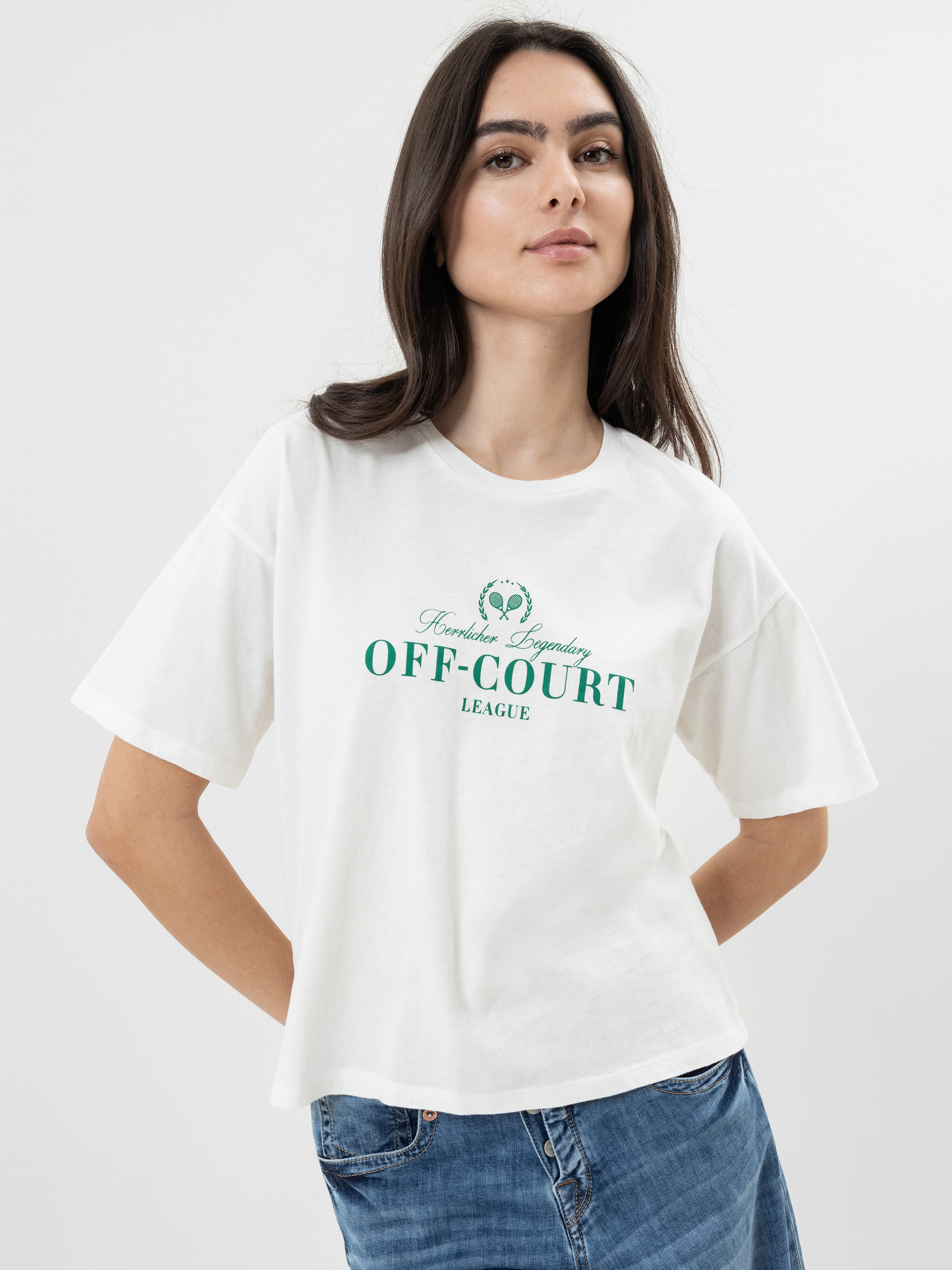 Eine Frau mit langen dunklen Haaren trägt ein lockeres weißes T-Shirt mit der grünen Aufschrift OFF-COURT LEAGUE und blaue Jeans. Sie steht mit einer Hand an der Hüfte vor einem schlichten hellen Hintergrund.
