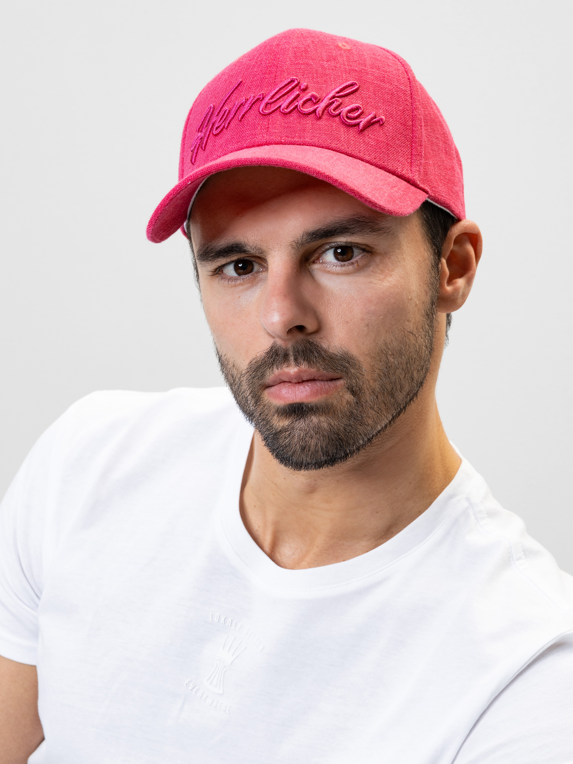 Ein Mann mit gestutztem Bart und Schnurrbart, der eine hellrosa Baseballkappe und ein schlichtes weißes T-Shirt trägt und vor einem schlichten hellgrauen Hintergrund posiert.