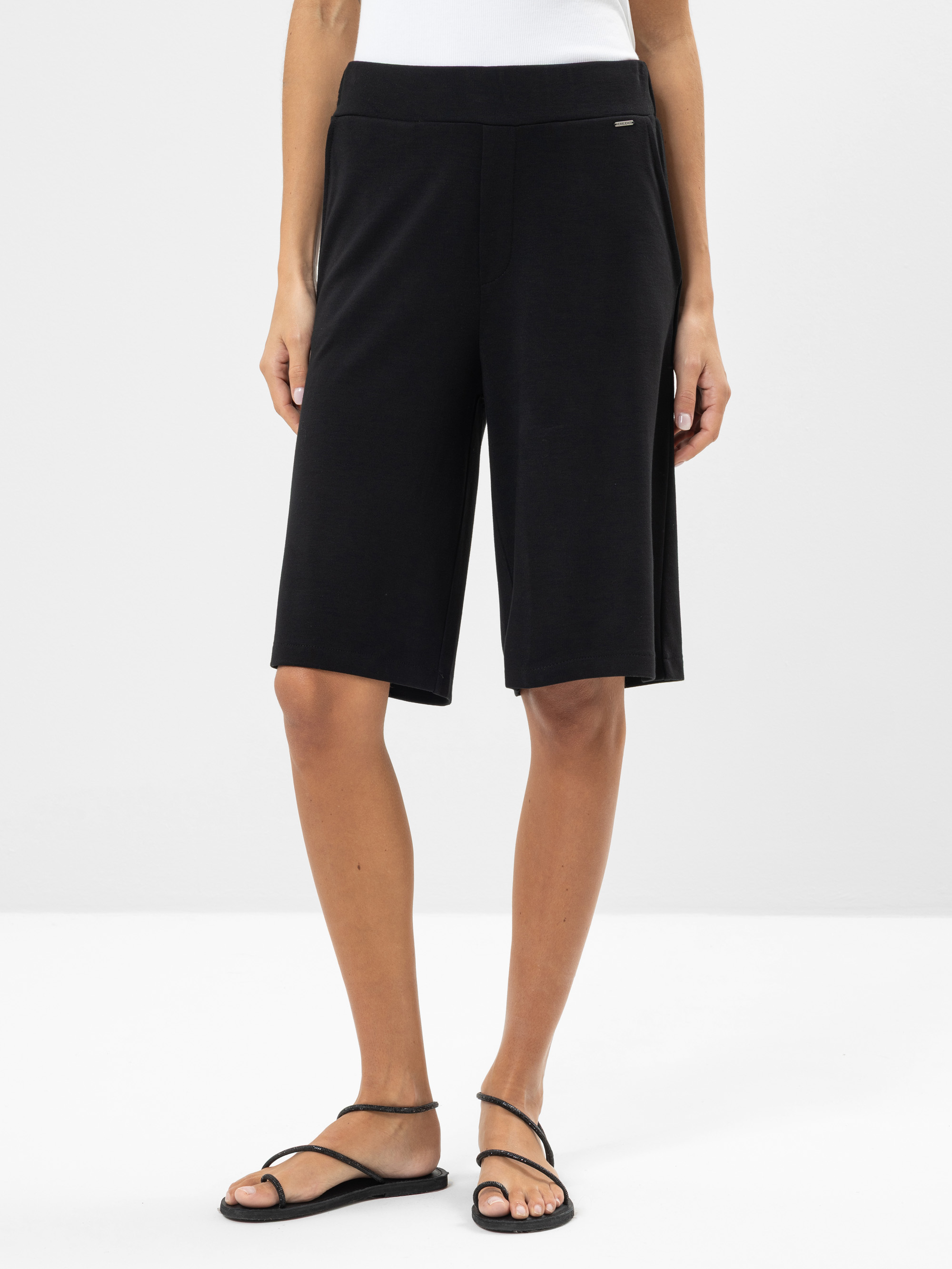Eine Person, die ein weißes Oberteil, locker sitzende knielange schwarze Shorts und schwarze Riemchensandalen trägt, steht vor einem schlichten hellen Hintergrund.