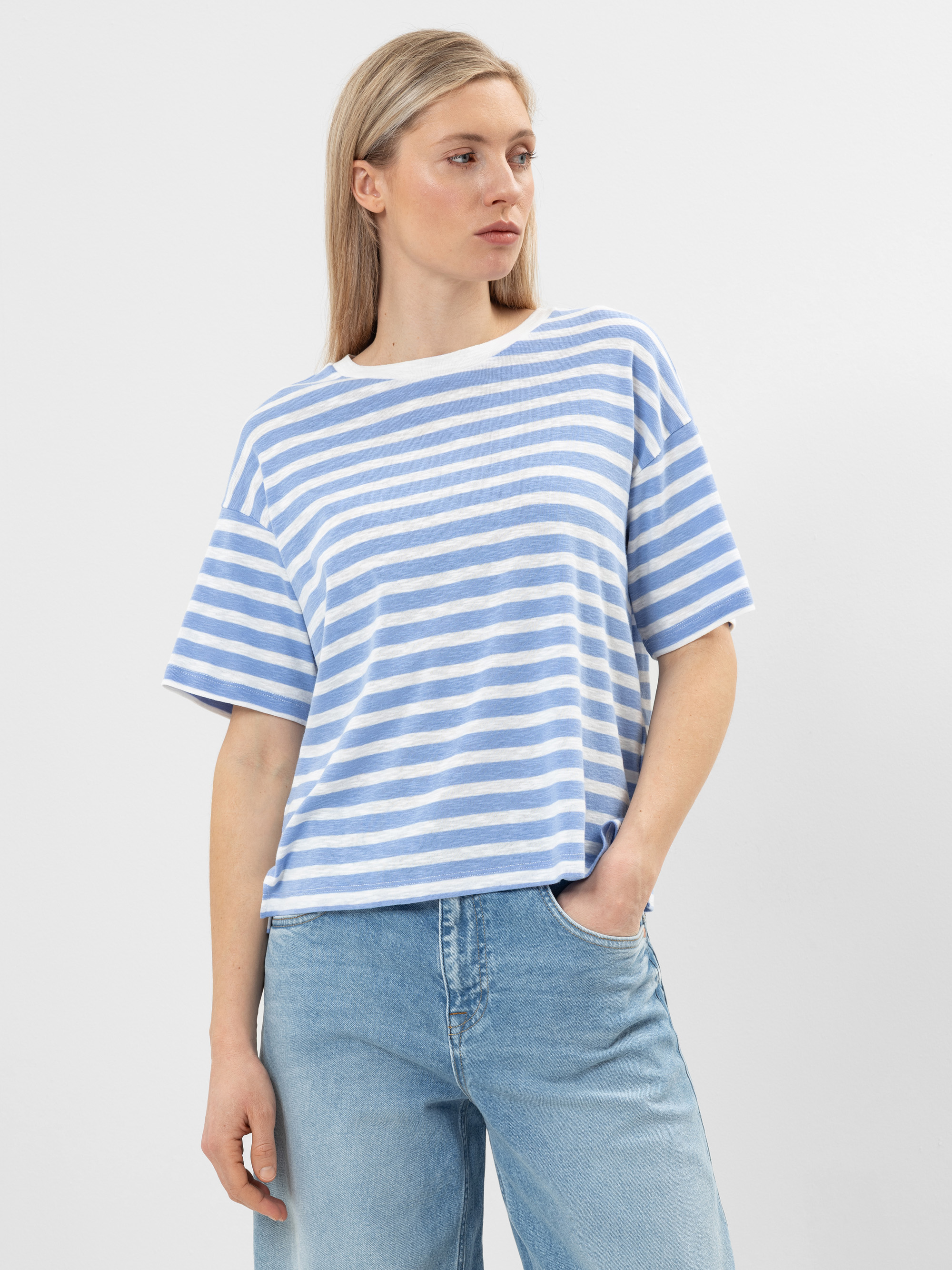 Eine Frau mit glattem blondem Haar trägt ein locker sitzendes, blau-weiß gestreiftes T-Shirt und hellblaue Jeans. Sie steht vor einem schlichten weißen Hintergrund und hat eine Hand in der Tasche.
