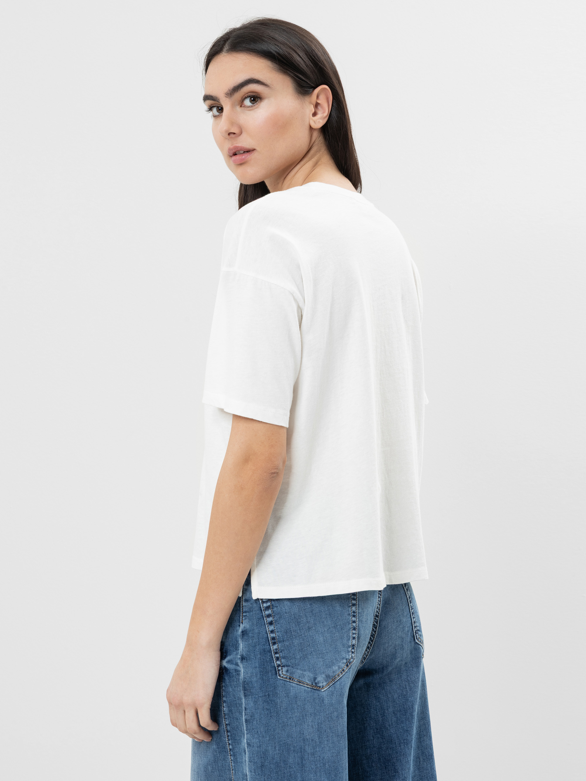 Eine Frau mit glattem dunklem Haar trägt ein lockeres weißes T-Shirt und eine blaue Jeans. Sie steht seitlich und schaut über ihre Schulter vor einem schlichten, hellen Hintergrund.