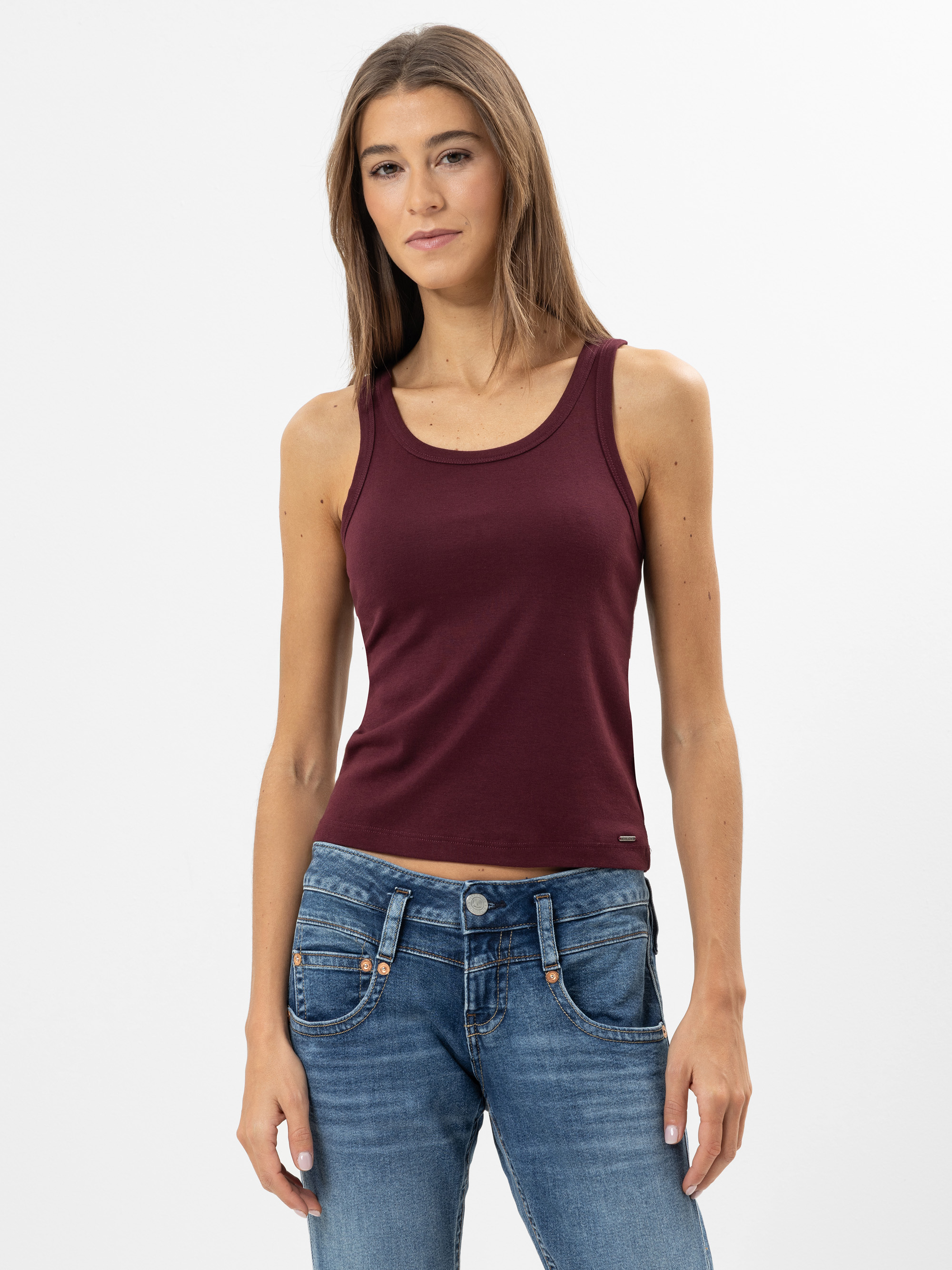 Eine Frau mit glattem braunem Haar trägt ein ärmelloses weinrotes Tanktop und blaue Jeans und steht vor einem schlichten weißen Hintergrund. Sie schaut mit neutralem Blick in die Kamera.