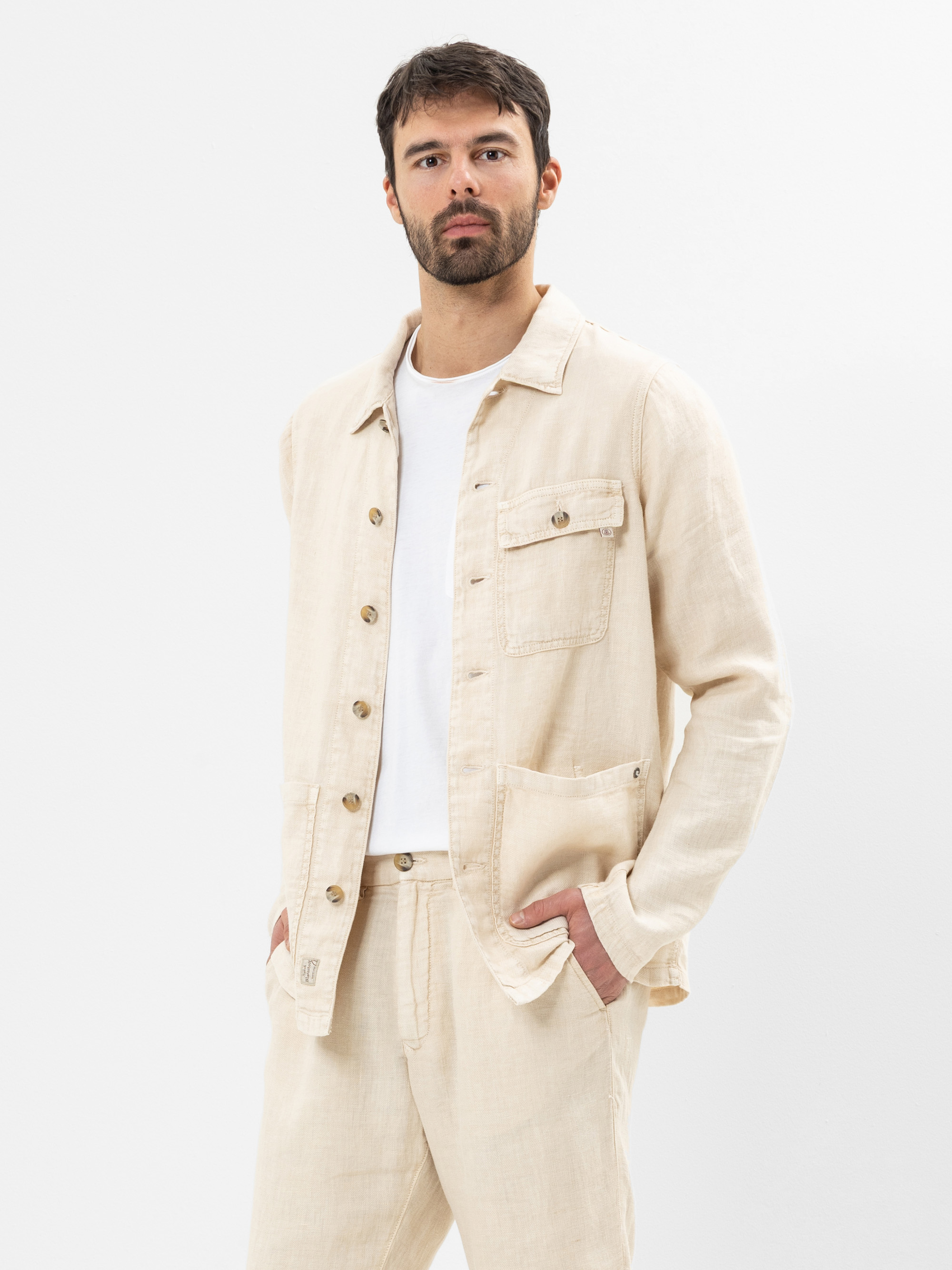 Ein Mann mit dunklem Haar und Bart trägt eine hellbeige Button-up-Hemdjacke über einem weißen T-Shirt und eine dazu passende beigefarbene Hose und steht mit einer Hand in der Tasche vor einem schlichten weißen Hintergrund.