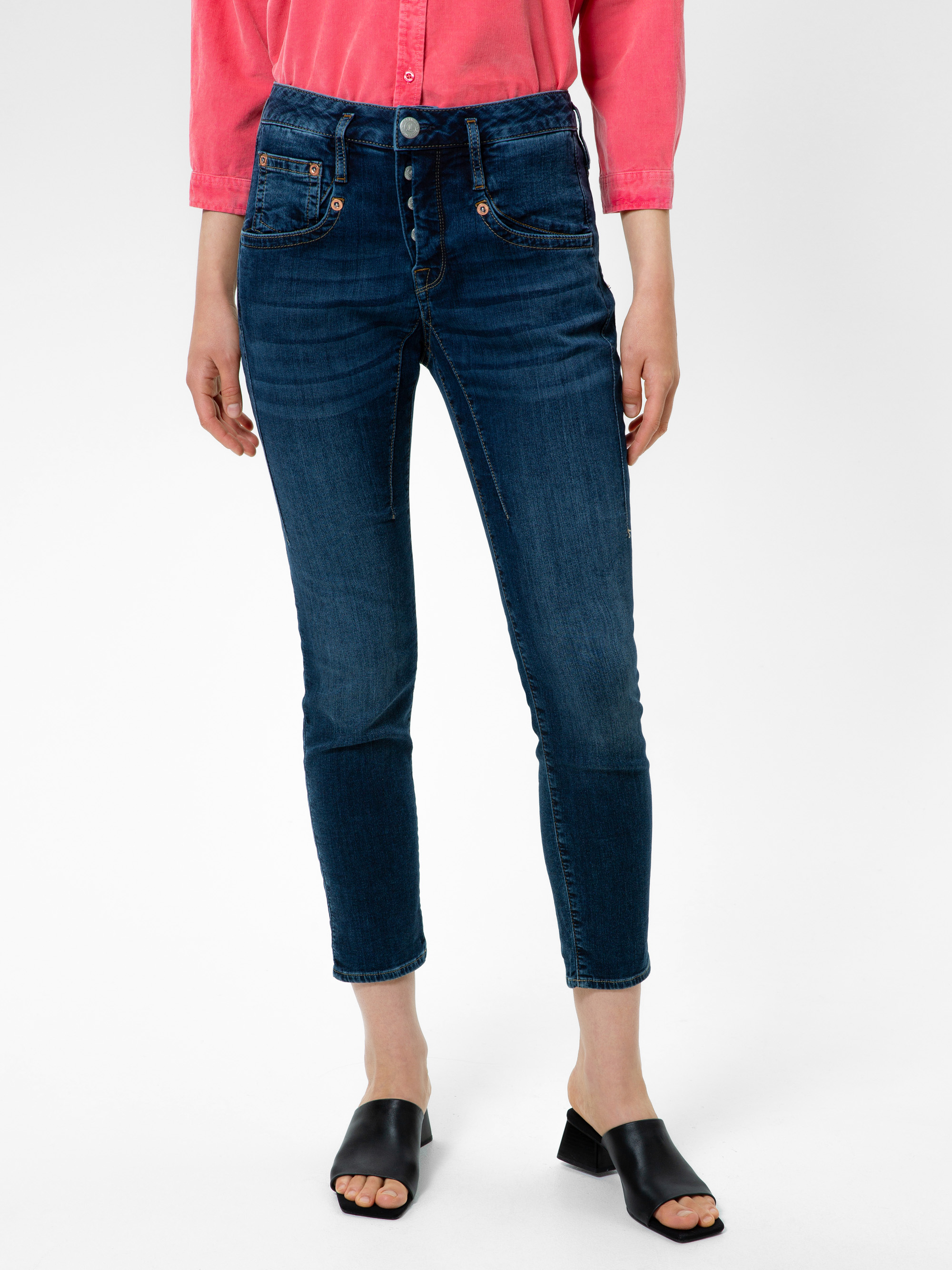 Eine Person, die ein korallenrotes Button-up-Hemd, hochgekrempelte dunkelblaue Skinny-Jeans und schwarze, offene Slipper-Sandalen trägt, steht vor einem schlichten weißen Hintergrund. Ihr Kopf ist auf dem Bild nicht zu sehen.