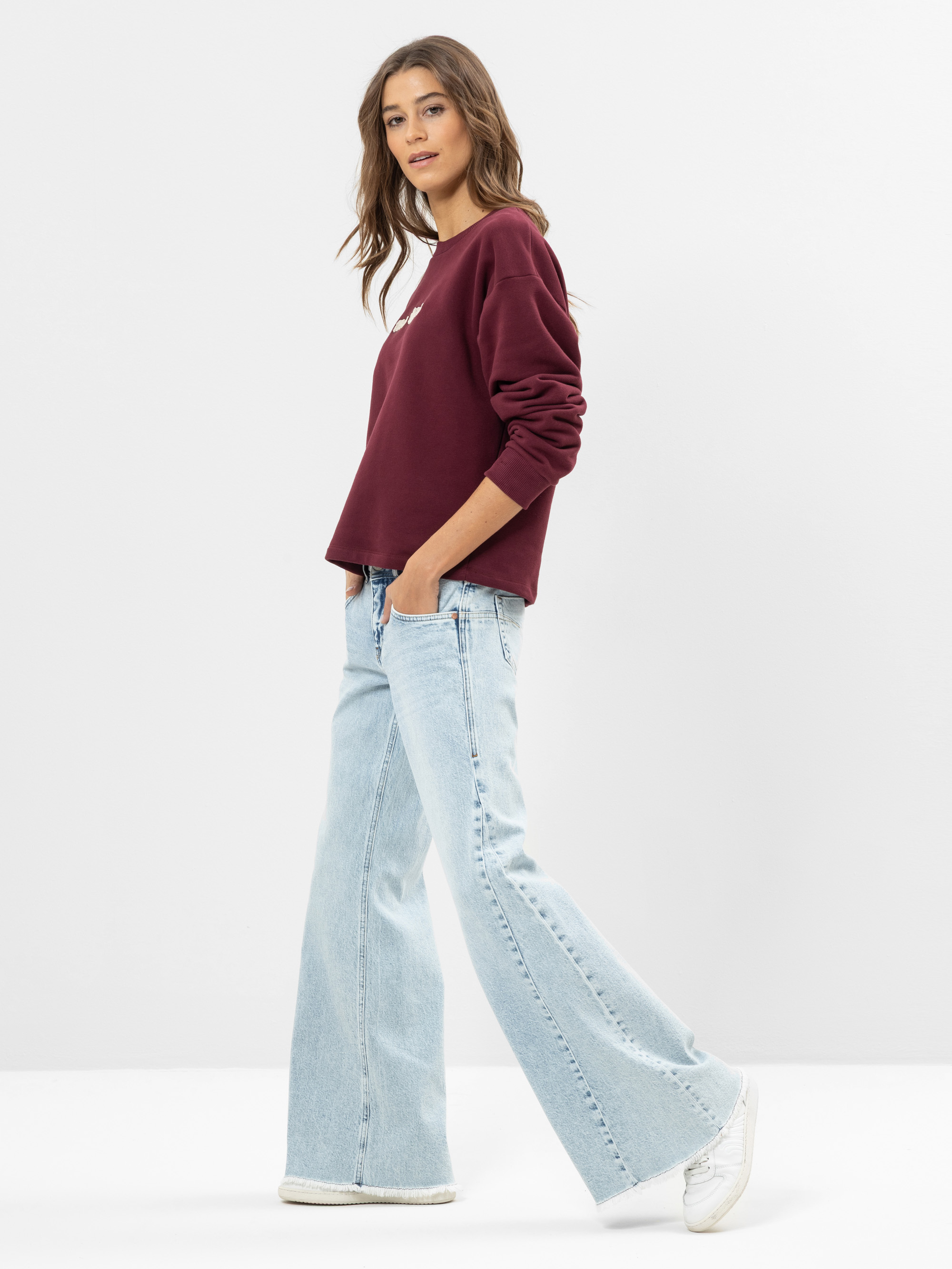 Eine Frau steht seitlich und trägt ein kastanienbraunes langärmeliges Oberteil, hellblaue Jeans mit weitem Bein und weiße Turnschuhe. Sie hat gewelltes braunes Haar und eine entspannte, lässige Pose vor einem schlichten weißen Hintergrund.