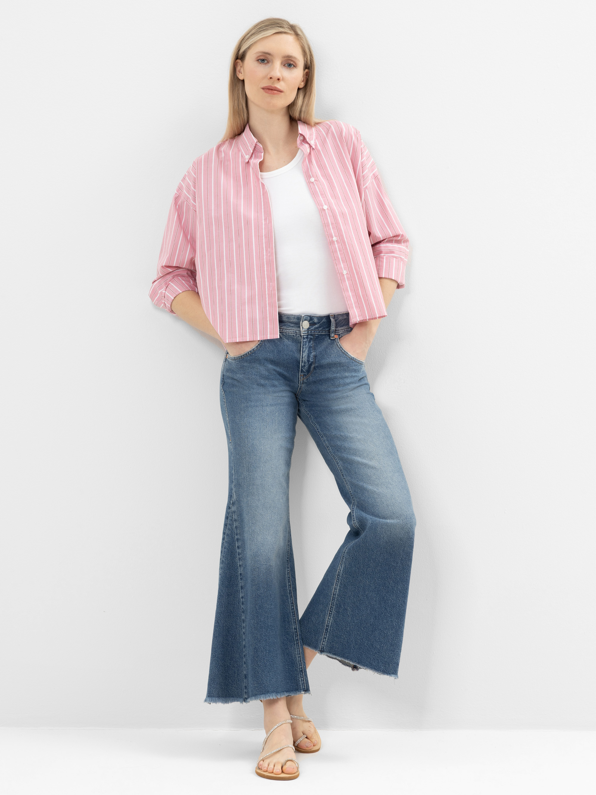 Eine Frau steht vor einer weißen Wand und trägt ein rosa-weiß gestreiftes Button-up-Hemd über einem weißen Oberteil, blaue Jeans mit weitem Bein und beigefarbene Sandalen. Sie hat glattes blondes Haar und eine entspannte Pose mit einer Hand in der Ta