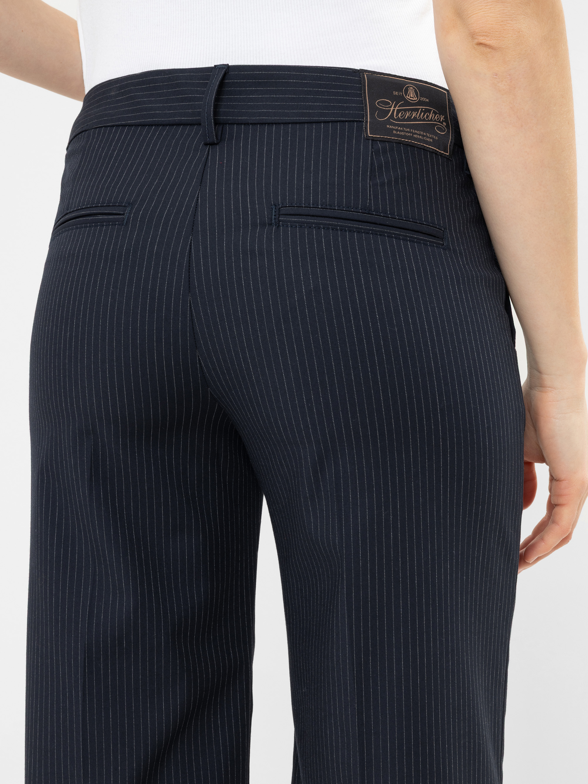 Eine Person trägt eine marineblaue Nadelstreifenhose und ein weißes Oberteil, von hinten gesehen. Die Hose hat zwei Gesäßtaschen und ein dunkles Etikett am Bund.