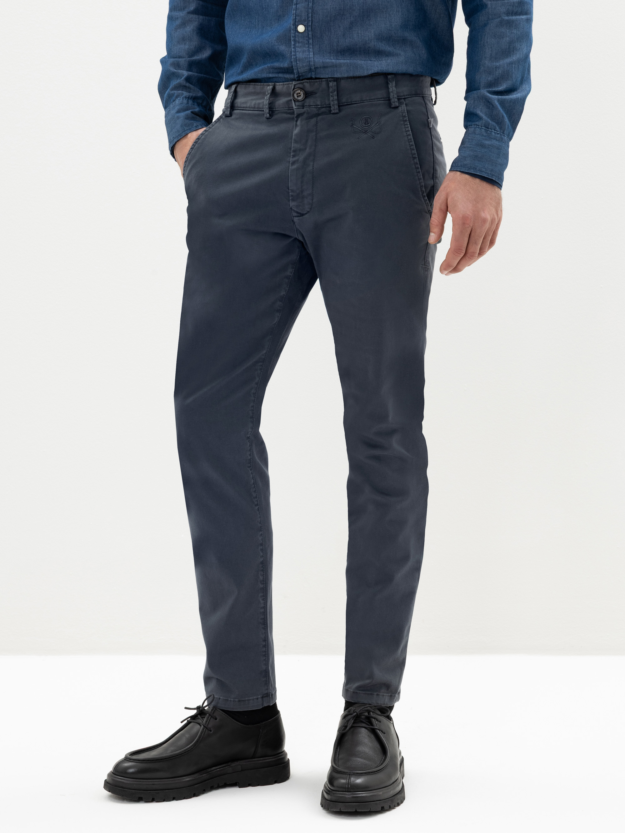 Ein Mann trägt eine dunkelblaue, maßgeschneiderte Chinohose, ein blaues Jeanshemd zum Umschlagen und schwarze Lederschuhe. Er steht mit den Händen in den Taschen vor einem schlichten weißen Hintergrund.