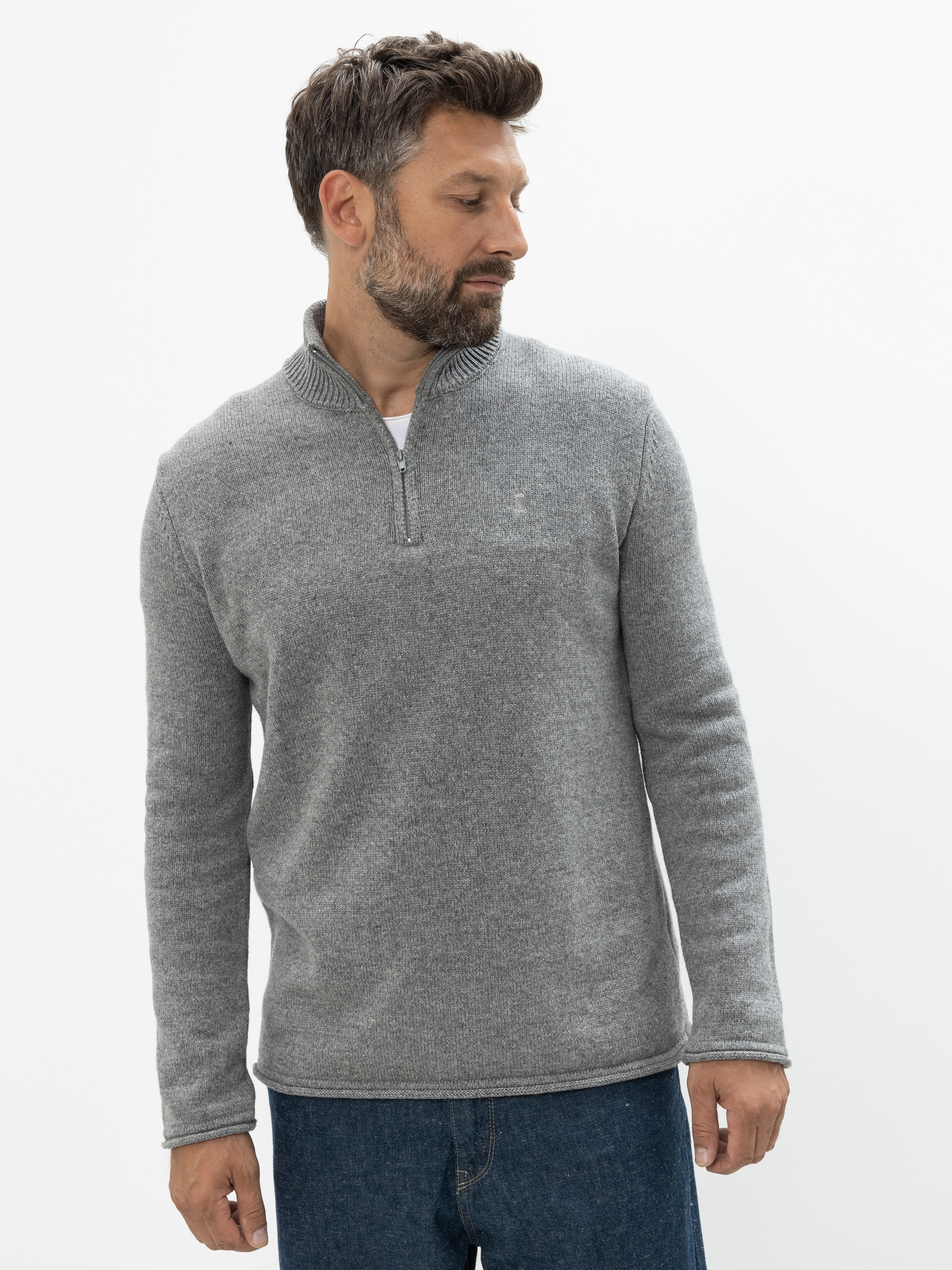 Ein Mann mit kurzen braunen Haaren und Bart trägt einen hellgrauen Pullover mit Viertelreißverschluss und blaue Jeans. Er steht vor einem schlichten weißen Hintergrund und blickt nach links.