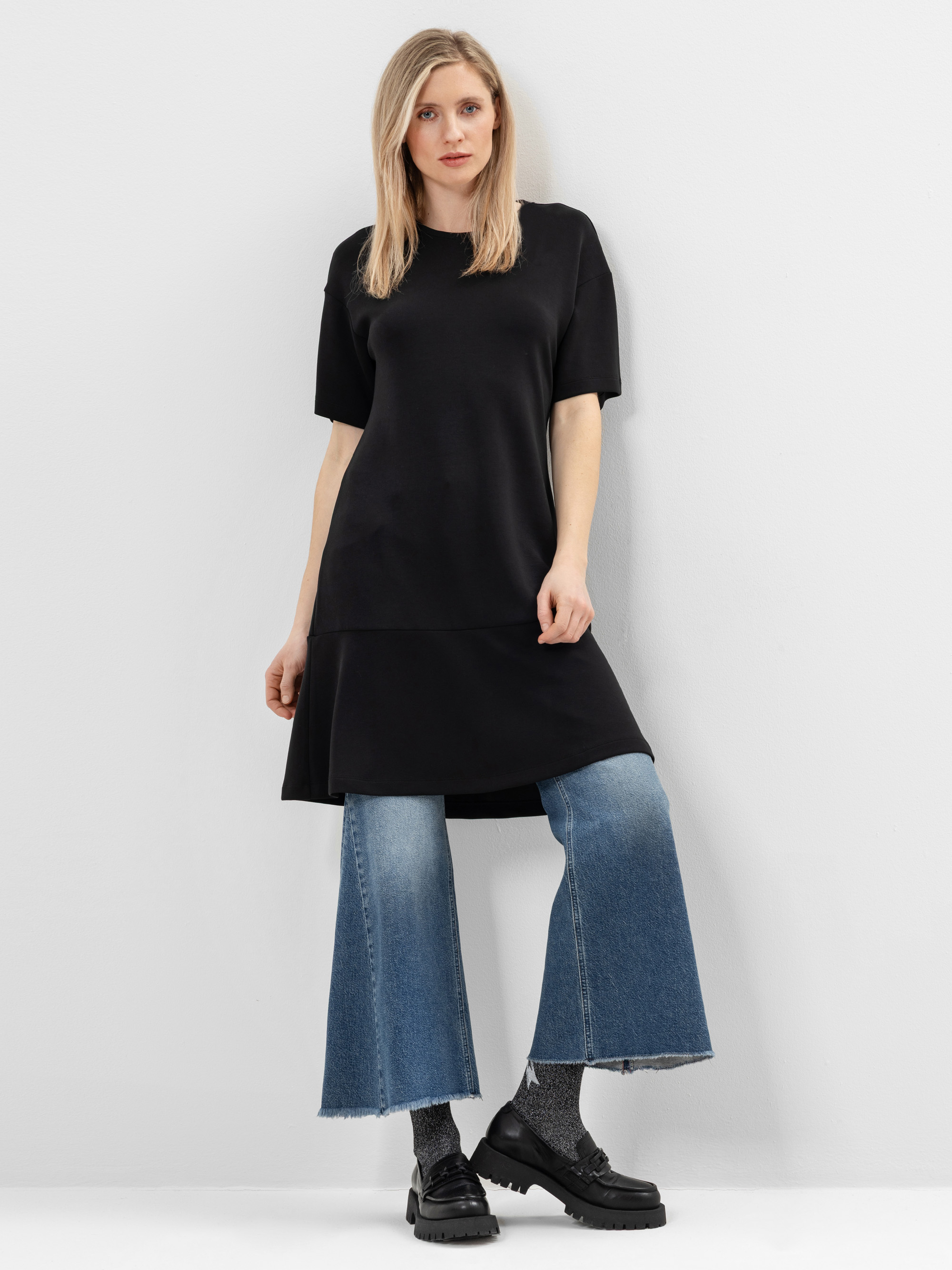 Eine Frau steht vor einer weißen Wand und trägt ein lockeres, schwarzes, kurzärmeliges Kleid über einer ausgestellten Bluejeans, dunkle Socken und schwarze Slipper. Sie hat glattes blondes Haar und einen neutralen Gesichtsausdruck.