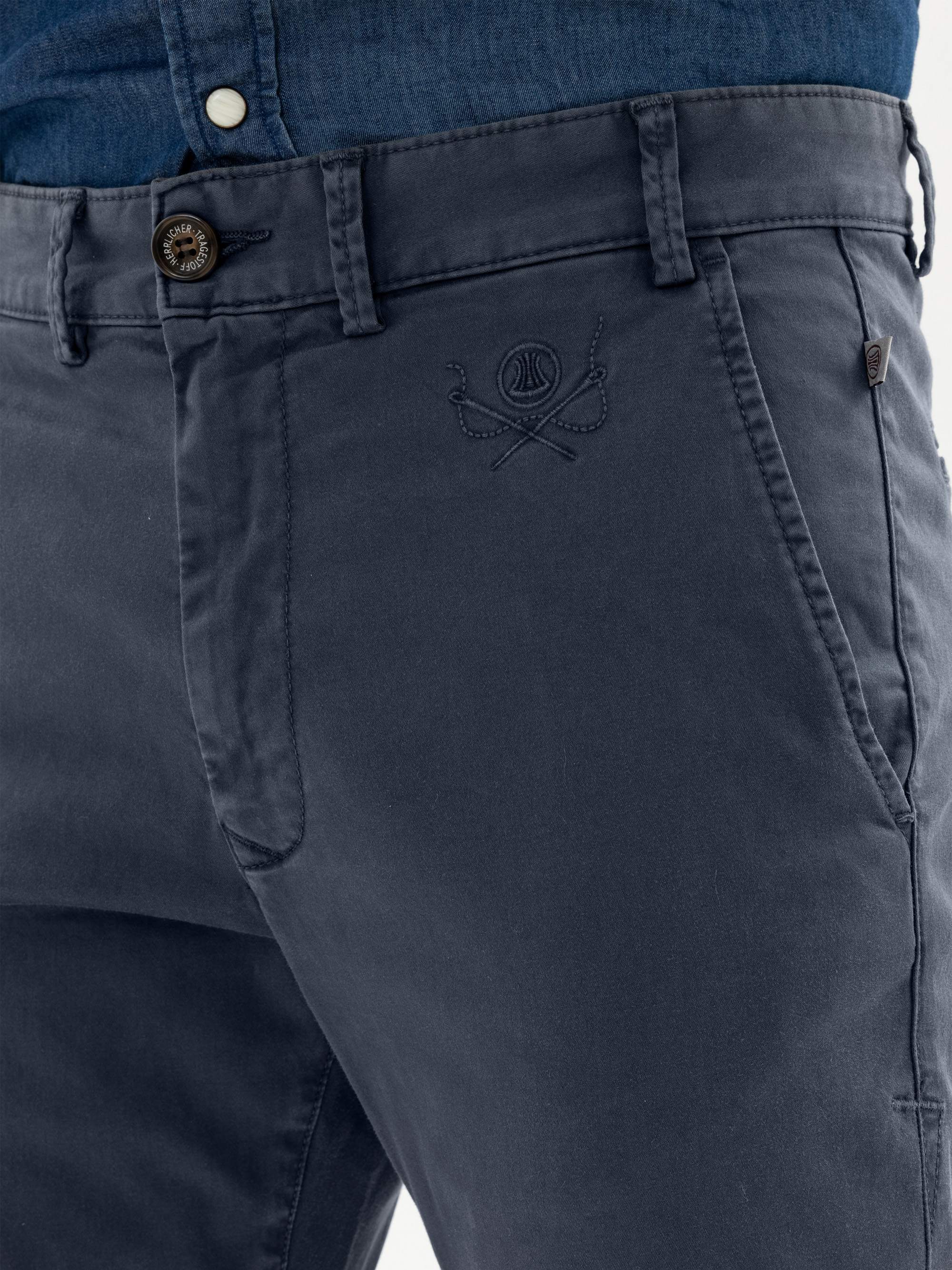 Nahaufnahme einer Person, die eine dunkelblaue Hose mit Knopf und Reißverschluss trägt, auf der ein gesticktes Logo über der rechten Tasche zu sehen ist, und ein in die Hose gestecktes Jeanshemd.