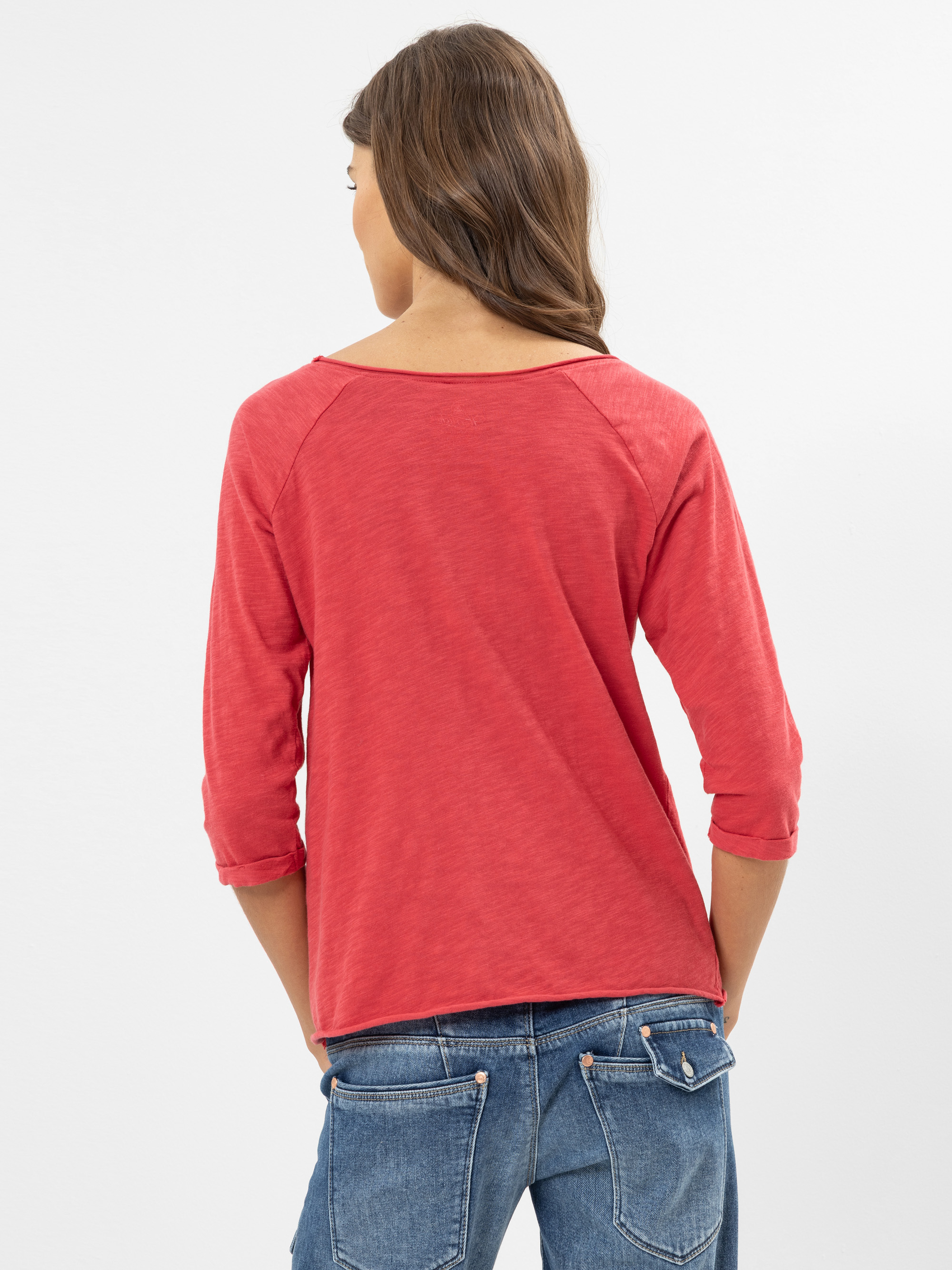 Eine Frau mit langen braunen Haaren steht mit dem Gesicht zur Wand, trägt ein rotes dreiviertelärmliges Oberteil und blaue Jeans mit Gesäßtaschen vor einem schlichten weißen Hintergrund.