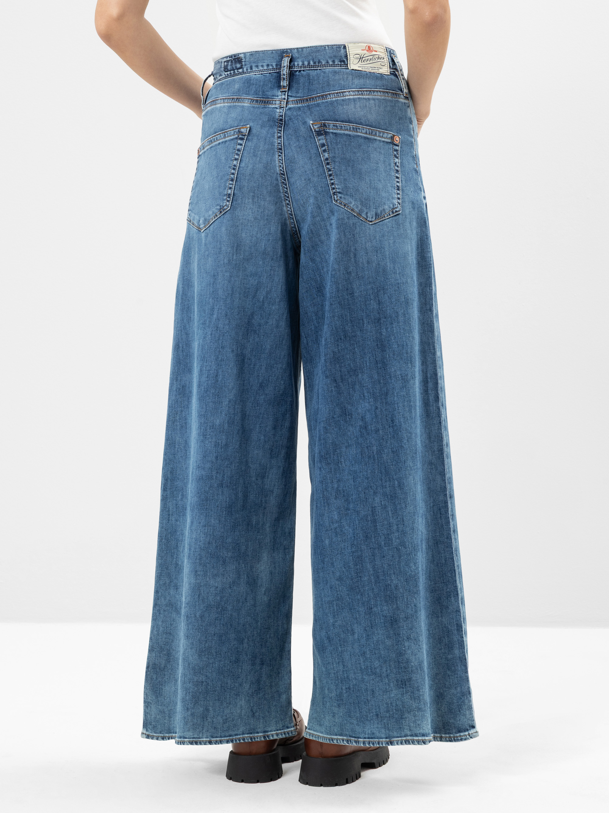 Eine Person, die eine Jeans mit weitem Bein und schwarze Schuhe trägt, wird von hinten gezeigt und steht vor einem schlichten weißen Hintergrund.