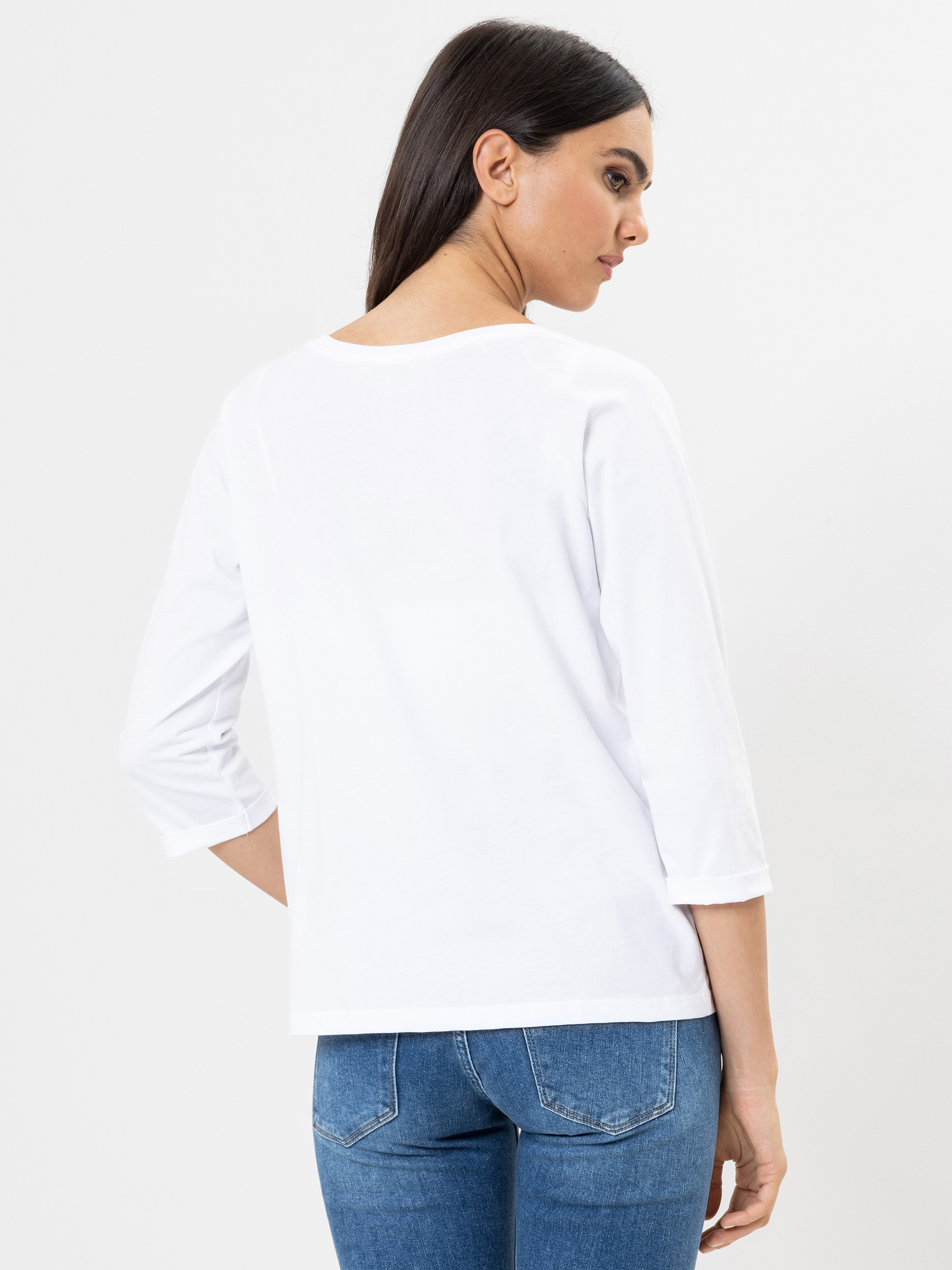 Eine Frau mit langen dunklen Haaren, die ein schlichtes weißes dreiviertelärmeliges Oberteil und blaue Jeans trägt, steht mit dem Gesicht zur Kamera vor einem weißen Hintergrund.