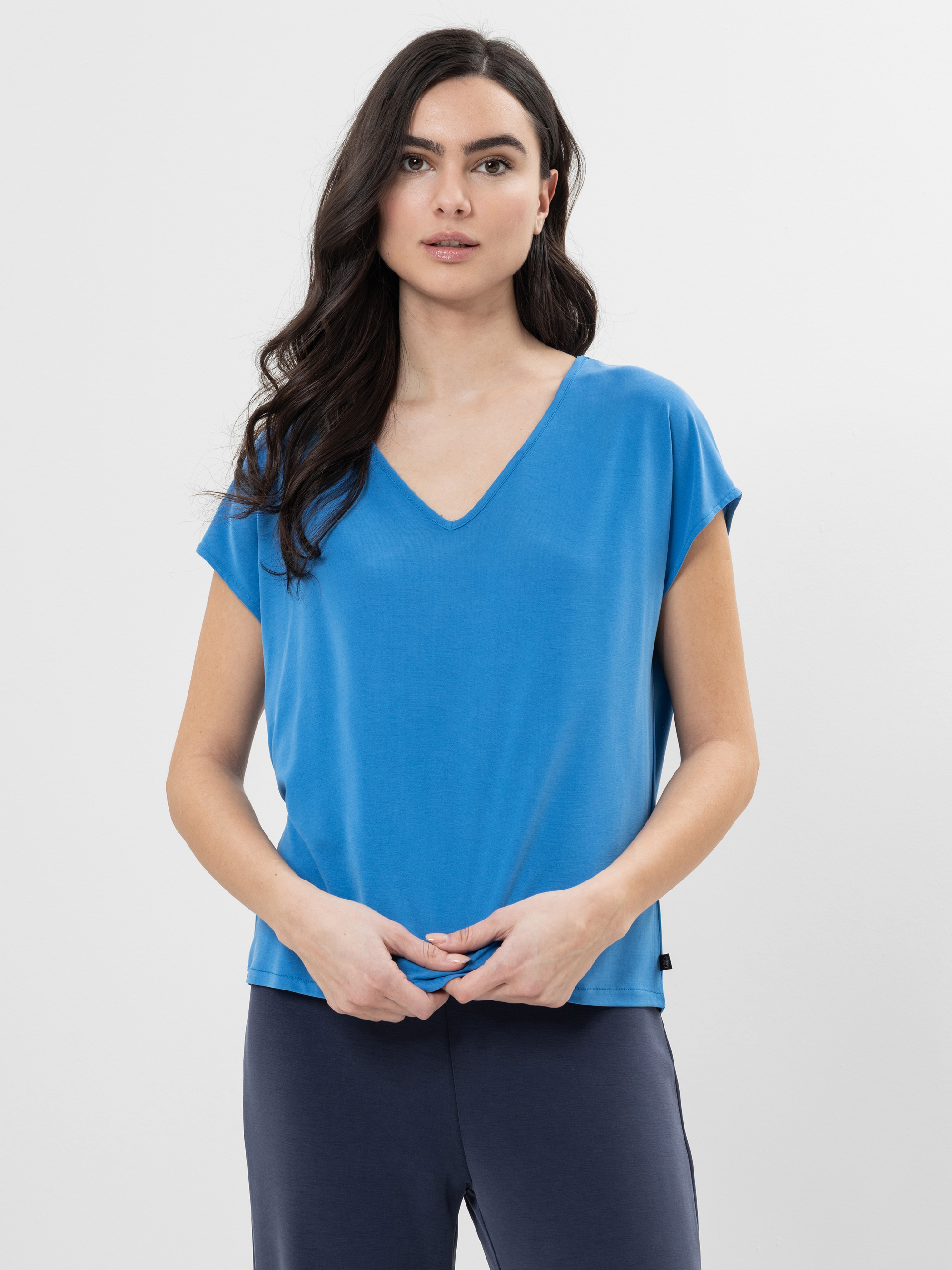 Eine Frau mit langen dunklen Haaren, die ein blaues kurzärmeliges Oberteil mit V-Ausschnitt und eine marineblaue Hose trägt, steht vor einem schlichten weißen Hintergrund und blickt nach vorne, wobei sie die Hände vor sich verschränkt.