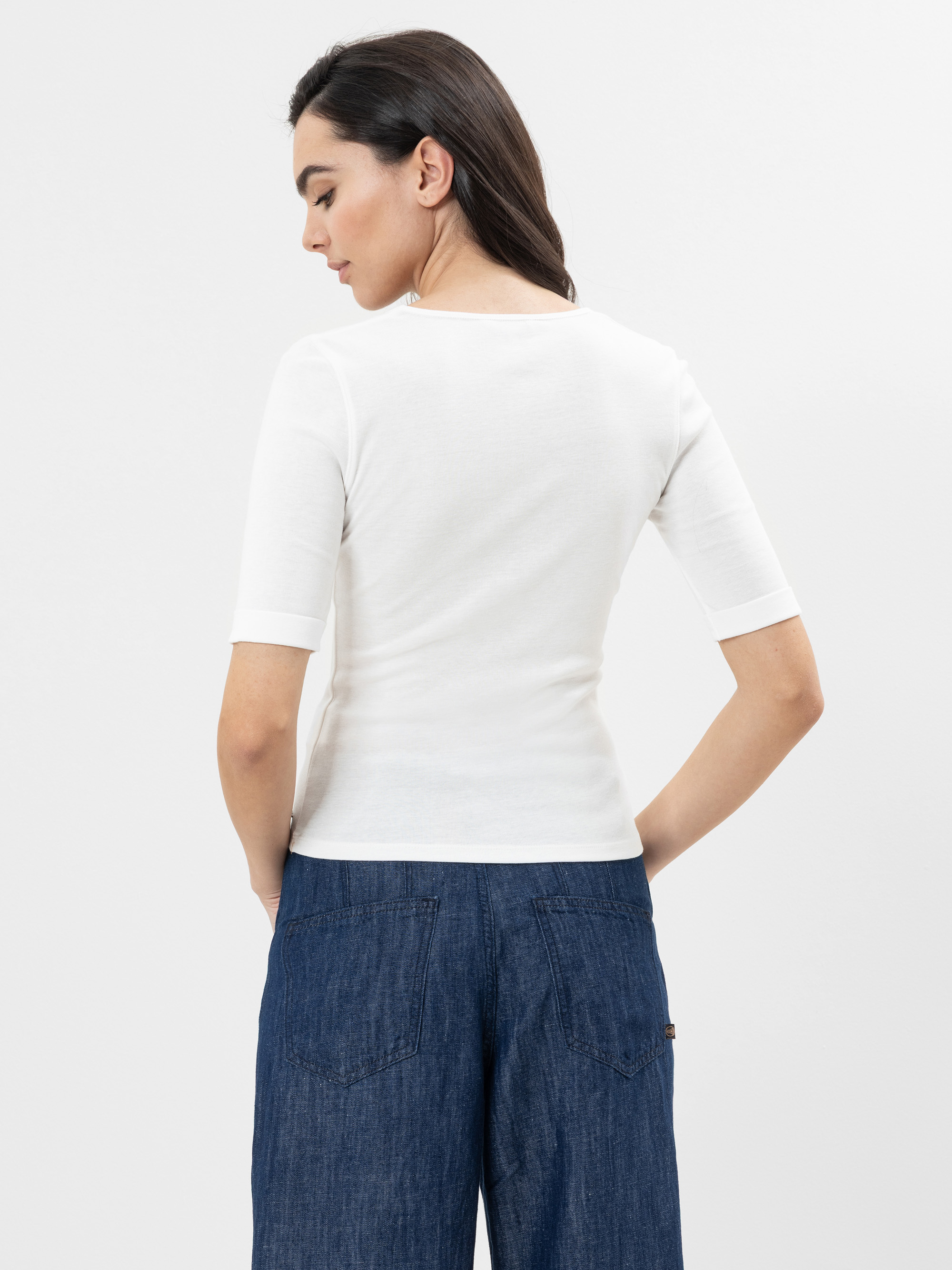 Frau steht mit dem Rücken zur Kamera, trägt ein weißes, kurzärmeliges Oberteil und eine blaue Jeanshose vor einem schlichten, hellen Hintergrund.