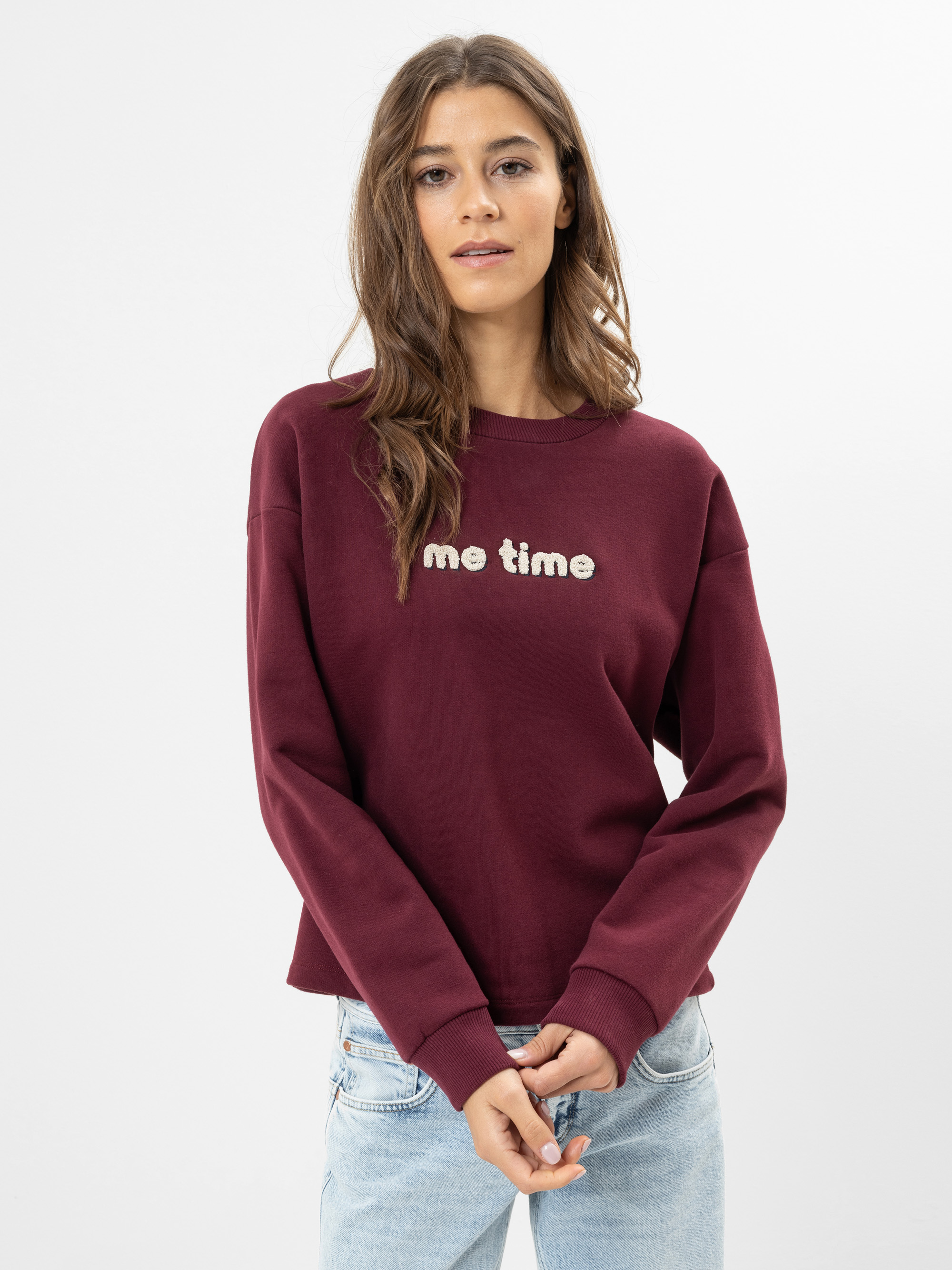 Eine Frau mit langen braunen Haaren, die ein kastanienbraunes Sweatshirt mit der Aufschrift me time trägt, steht vor einem schlichten weißen Hintergrund. Sie trägt außerdem eine hellblaue Jeans und hat die Hände locker vor sich verschränkt.