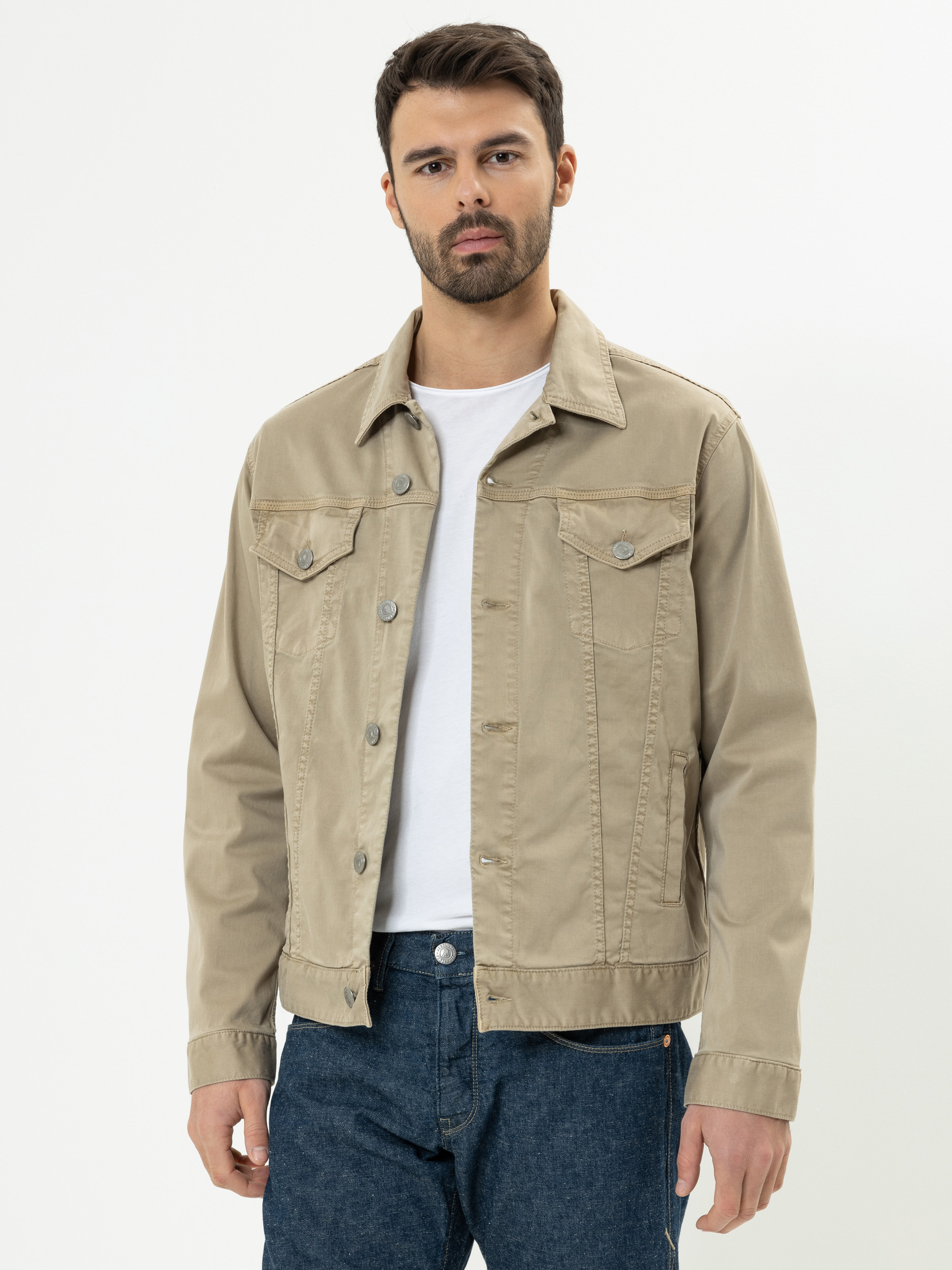 Ein Mann mit dunklem Haar und Bart trägt eine beige Jeansjacke über einem weißen T-Shirt und blaue Jeans und steht vor einem schlichten weißen Hintergrund.
