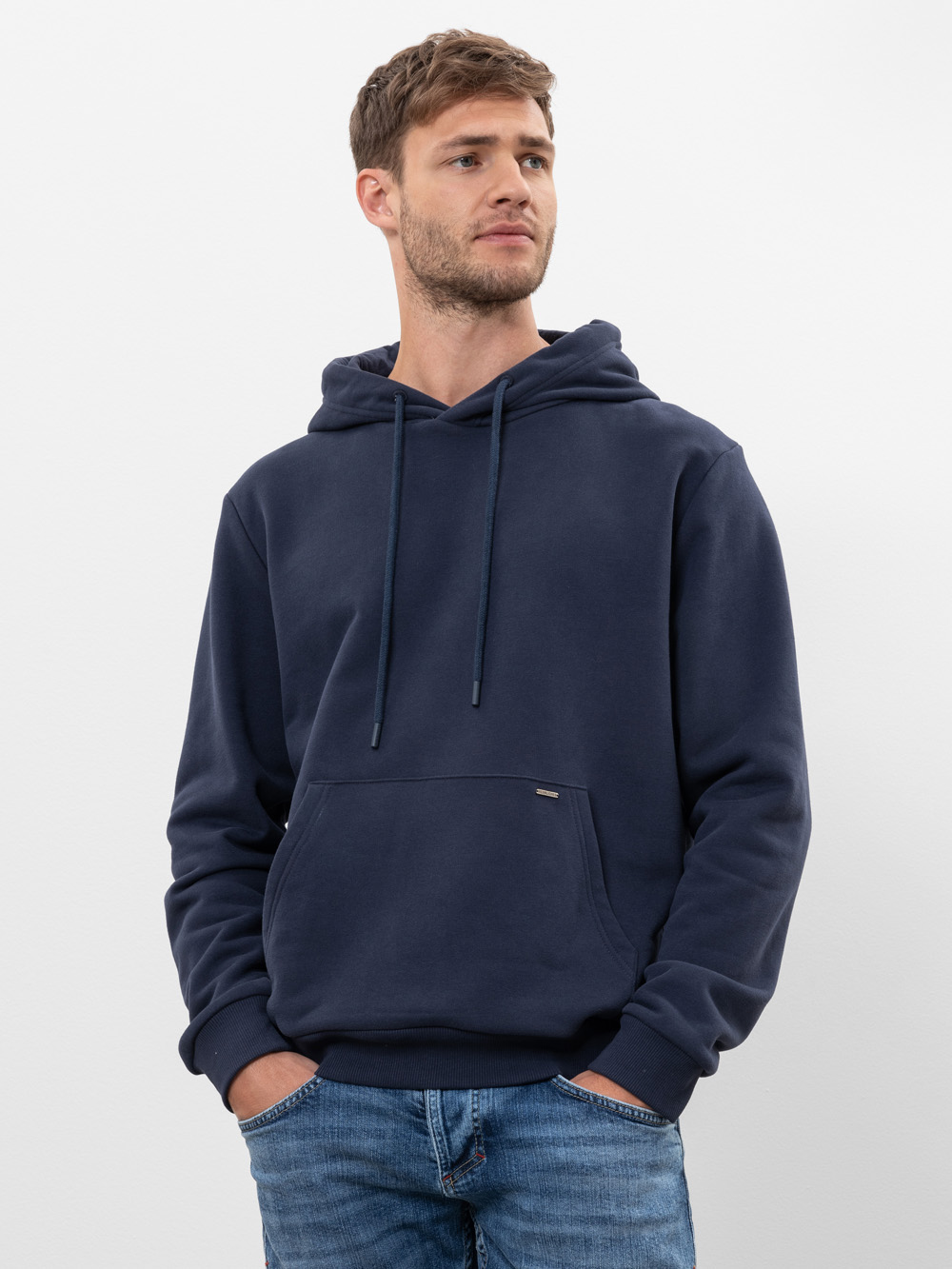 Ein Mann mit kurzen braunen Haaren und gestutztem Bart trägt einen dunkelblauen Kapuzenpullover mit einer Vordertasche und hellblaue Jeans und steht mit den Händen in den Taschen vor einem schlichten weißen Hintergrund.