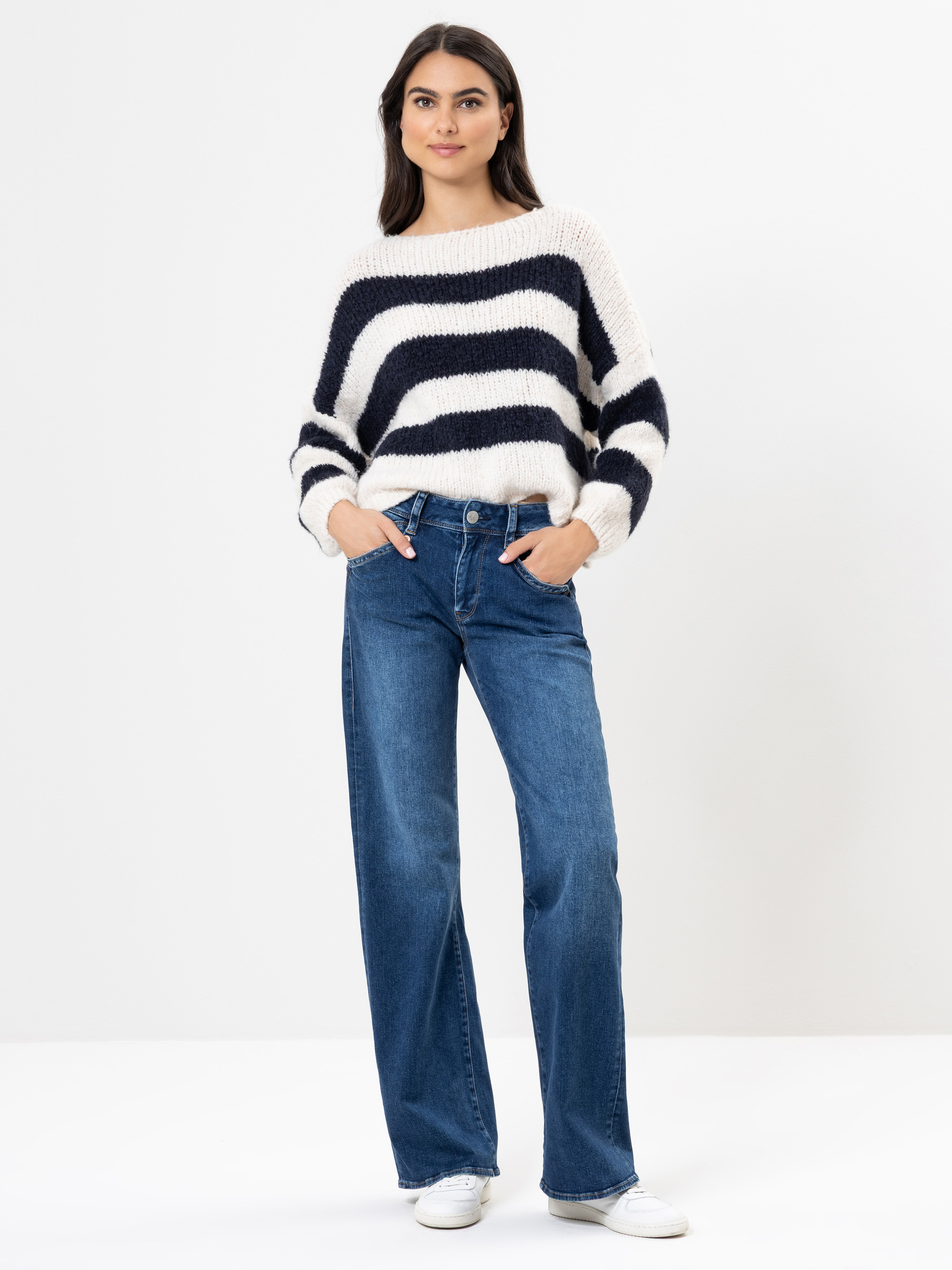 Eine Frau steht vor einem weißen Hintergrund und trägt einen wuscheligen, schwarz-weiß gestreiften Pullover, blaue Jeans mit weitem Bein und weiße Turnschuhe. Sie hat lange dunkle Haare und ihre Hände stecken in den Taschen.