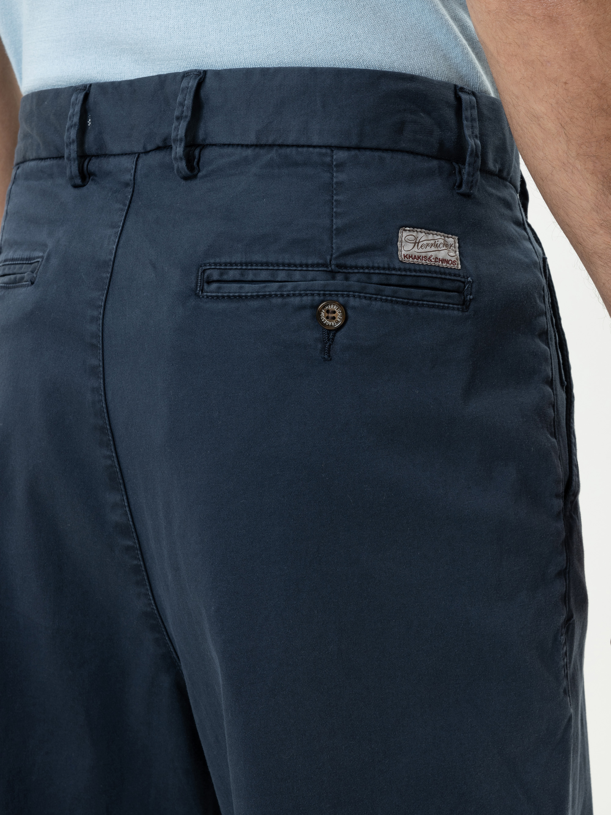 Nahaufnahme einer Person, die eine dunkelblaue Hose und ein hellblaues Hemd trägt. Der Fokus liegt auf der Gesäßtasche, die mit einem Knopf und einem kleinen rechteckigen Etikett mit Text in der Nähe des Hosenbundes versehen ist.