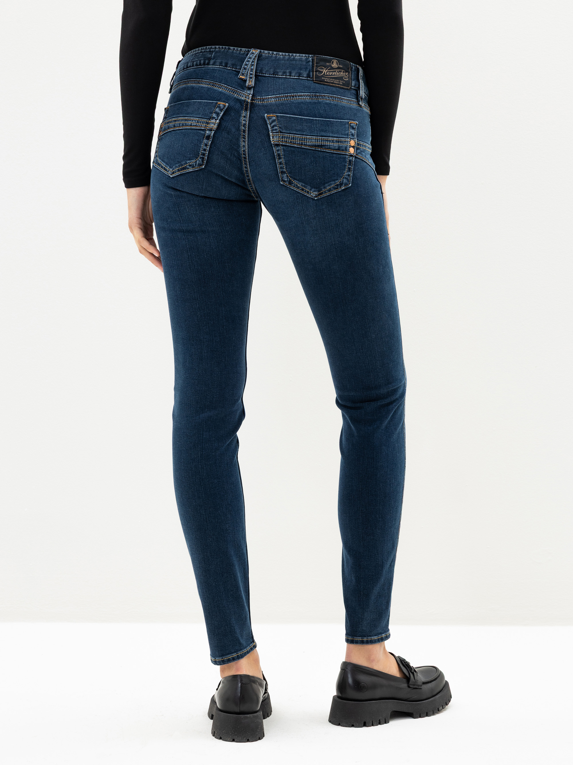 Eine Person, die dunkelblaue Röhrenjeans, ein schwarzes langärmeliges Oberteil und schwarze Halbschuhe trägt, steht mit dem Gesicht zur Kamera vor einem schlichten weißen Hintergrund.