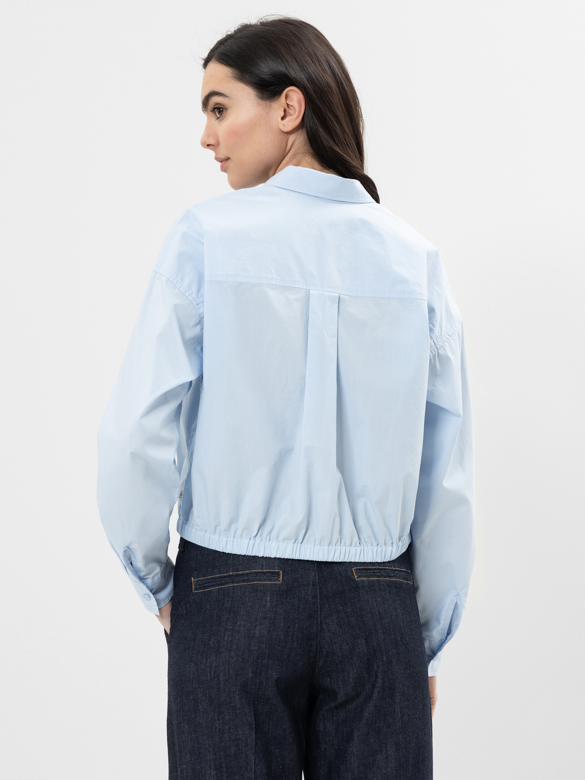 Eine Frau mit dunklem Haar trägt ein hellblaues Hemd mit Kragen und langen Ärmeln und dunkle Jeans mit hoher Taille. Sie schaut von sich weg und zeigt die Rückseite ihres Outfits vor einem schlichten weißen Hintergrund.