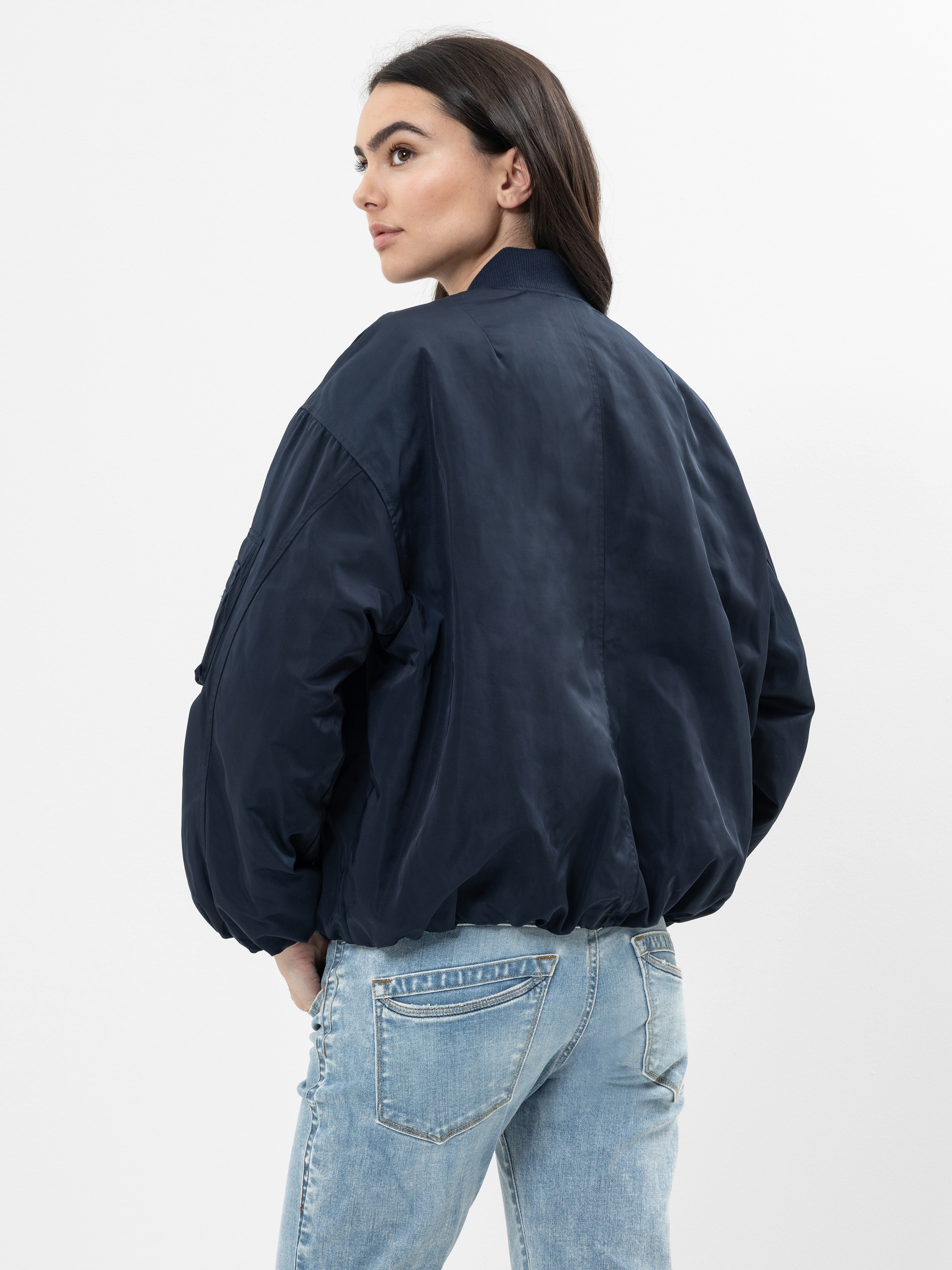 Eine Person mit langen dunklen Haaren trägt eine marineblaue Bomberjacke und hellblaue Jeans, steht vor einem schlichten weißen Hintergrund und blickt über ihre Schulter.