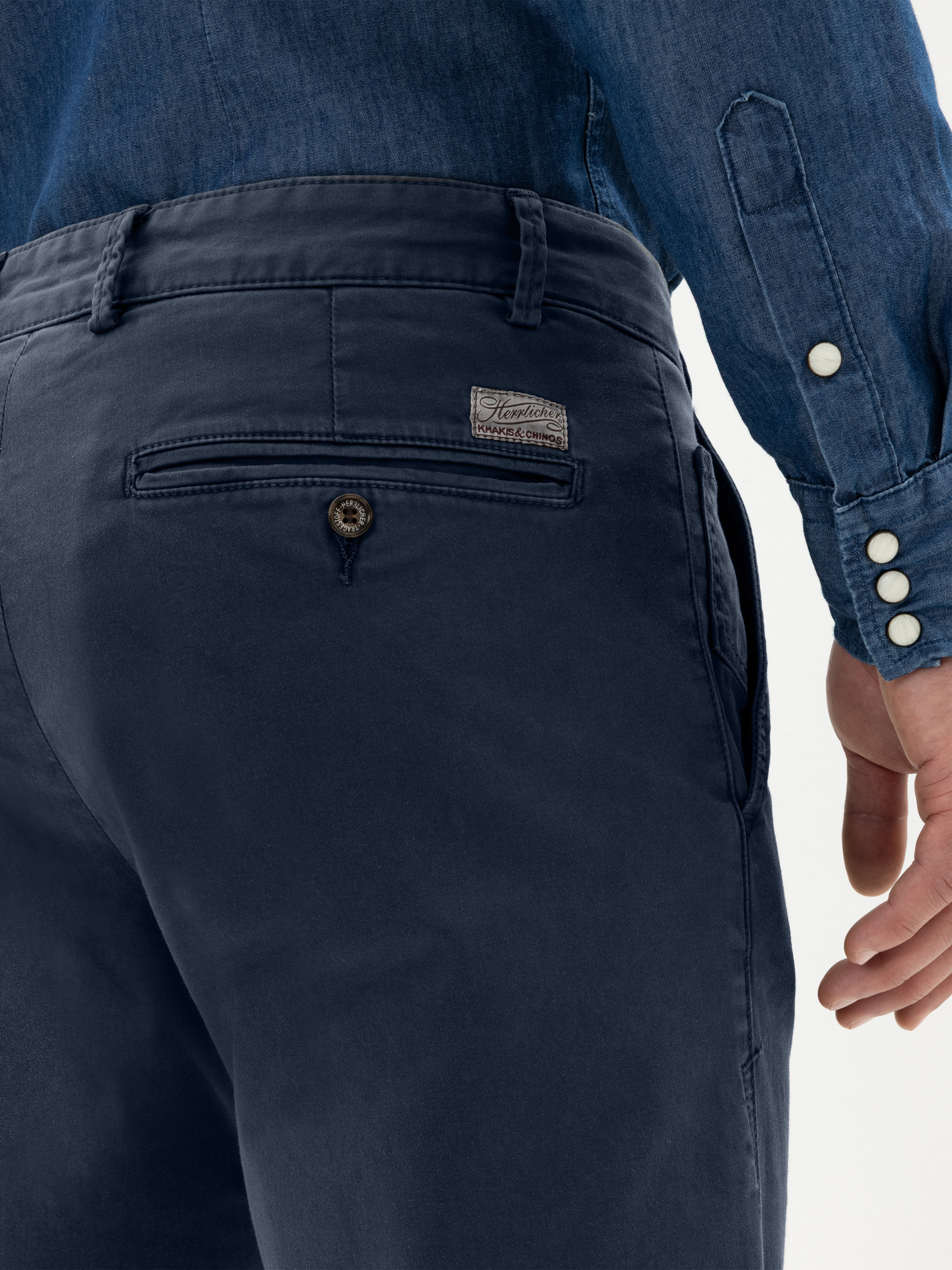 Nahaufnahme einer Person, die eine dunkelblaue Hose mit einer geknöpften Gesäßtasche und ein Jeanshemd mit weißen Knöpfen trägt. Die rechte Hand der Person ist an ihrer Seite zu sehen.
