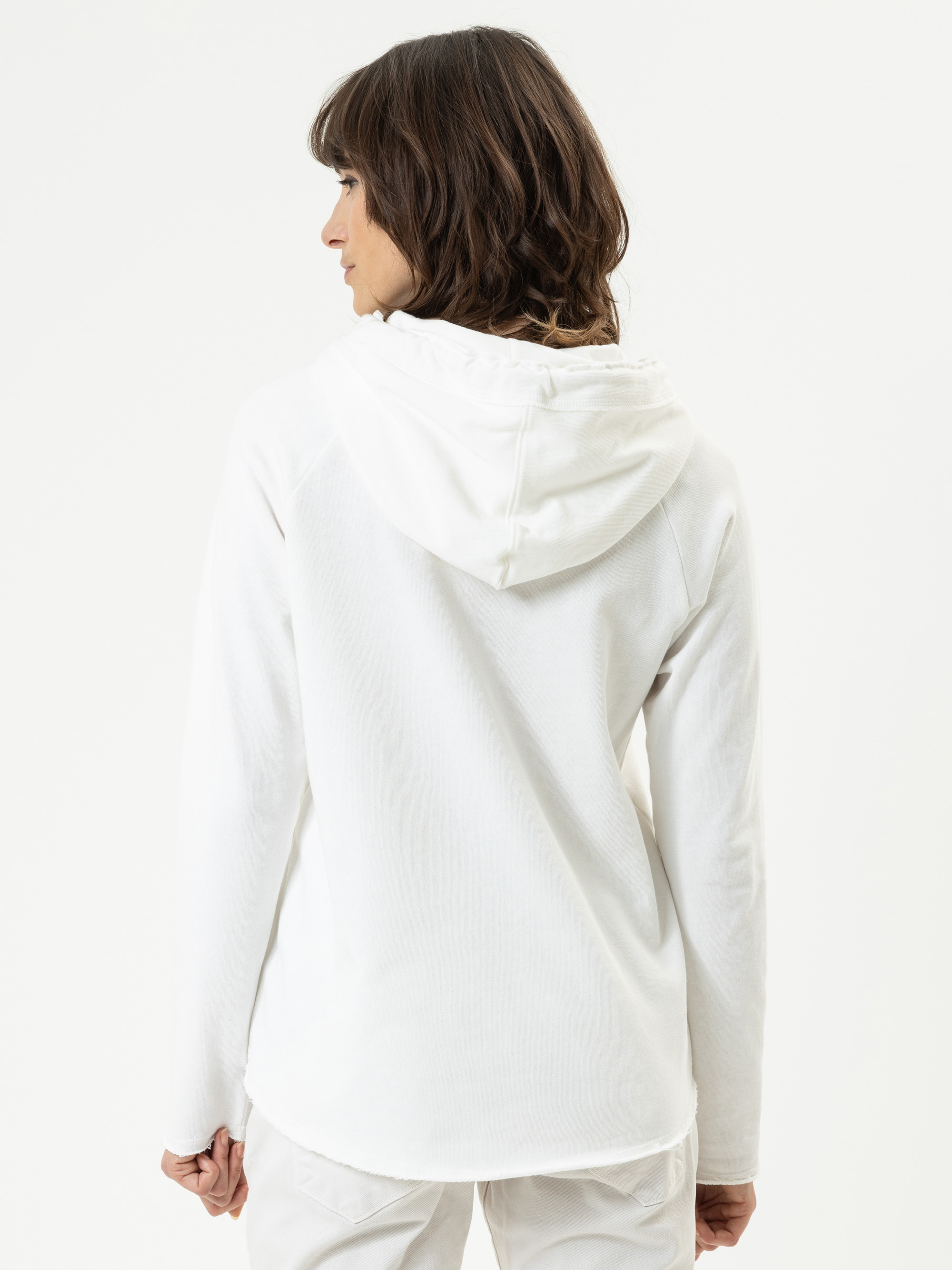 Eine Person mit schulterlangen braunen Haaren steht mit dem Gesicht zur Wand, trägt ein weißes Kapuzensweatshirt und eine weiße Hose vor einem schlichten weißen Hintergrund.