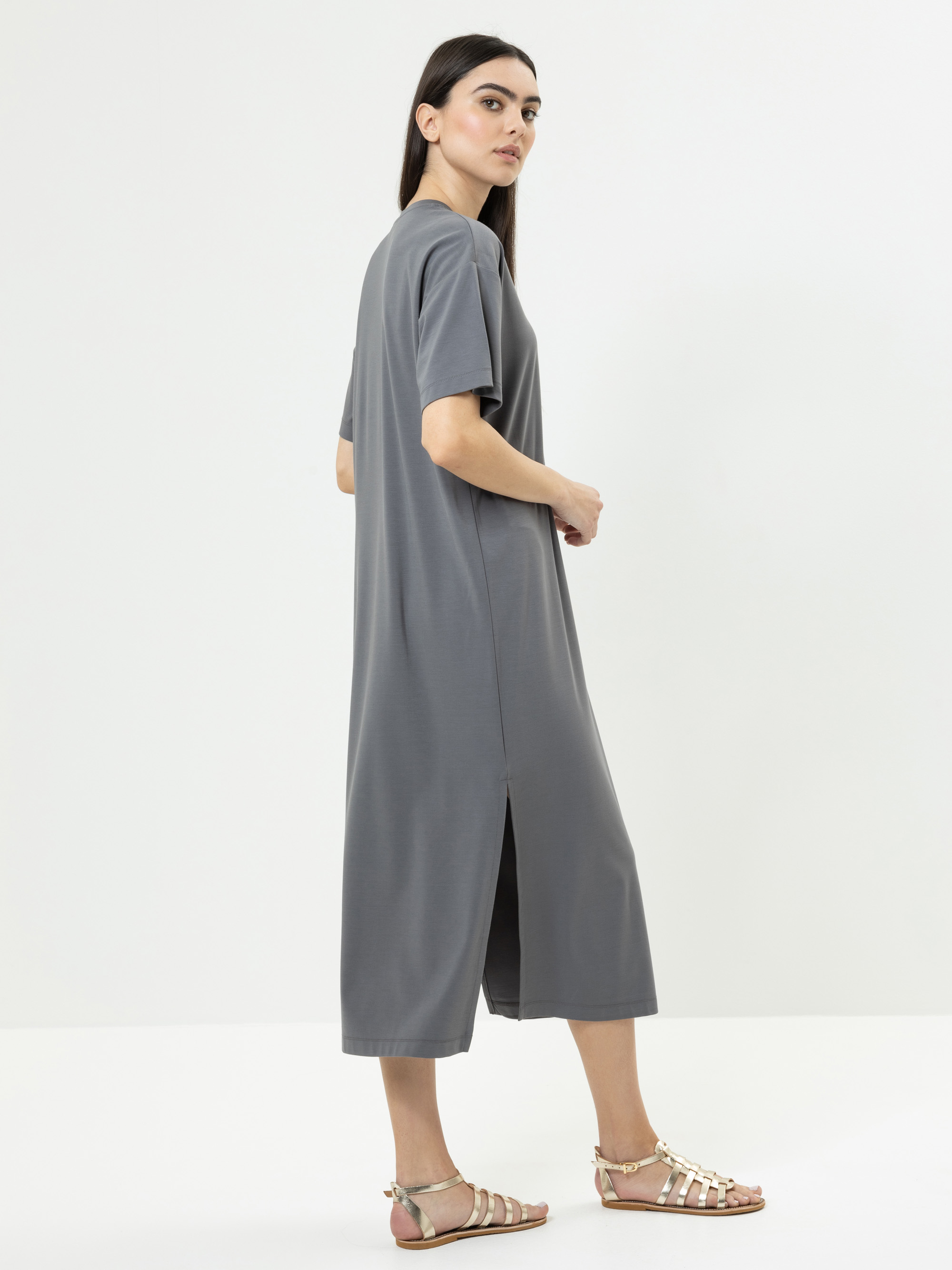 Eine Frau steht seitlich und trägt ein locker sitzendes, graues, kurzärmeliges Midikleid mit einem Seitenschlitz und flache Metallic-Sandalen vor einem schlichten weißen Hintergrund.