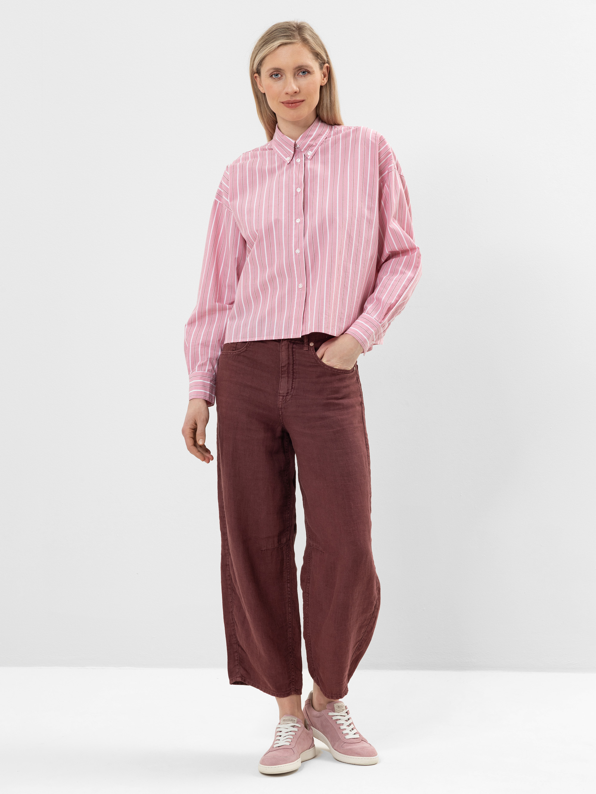 Eine Frau steht vor einem schlichten weißen Hintergrund und trägt ein rosa-weiß gestreiftes Button-up-Hemd, eine weit geschnittene kastanienbraune Hose und rosa Turnschuhe. Sie hat glattes blondes Haar und eine entspannte, lässige Pose mit einer Hand