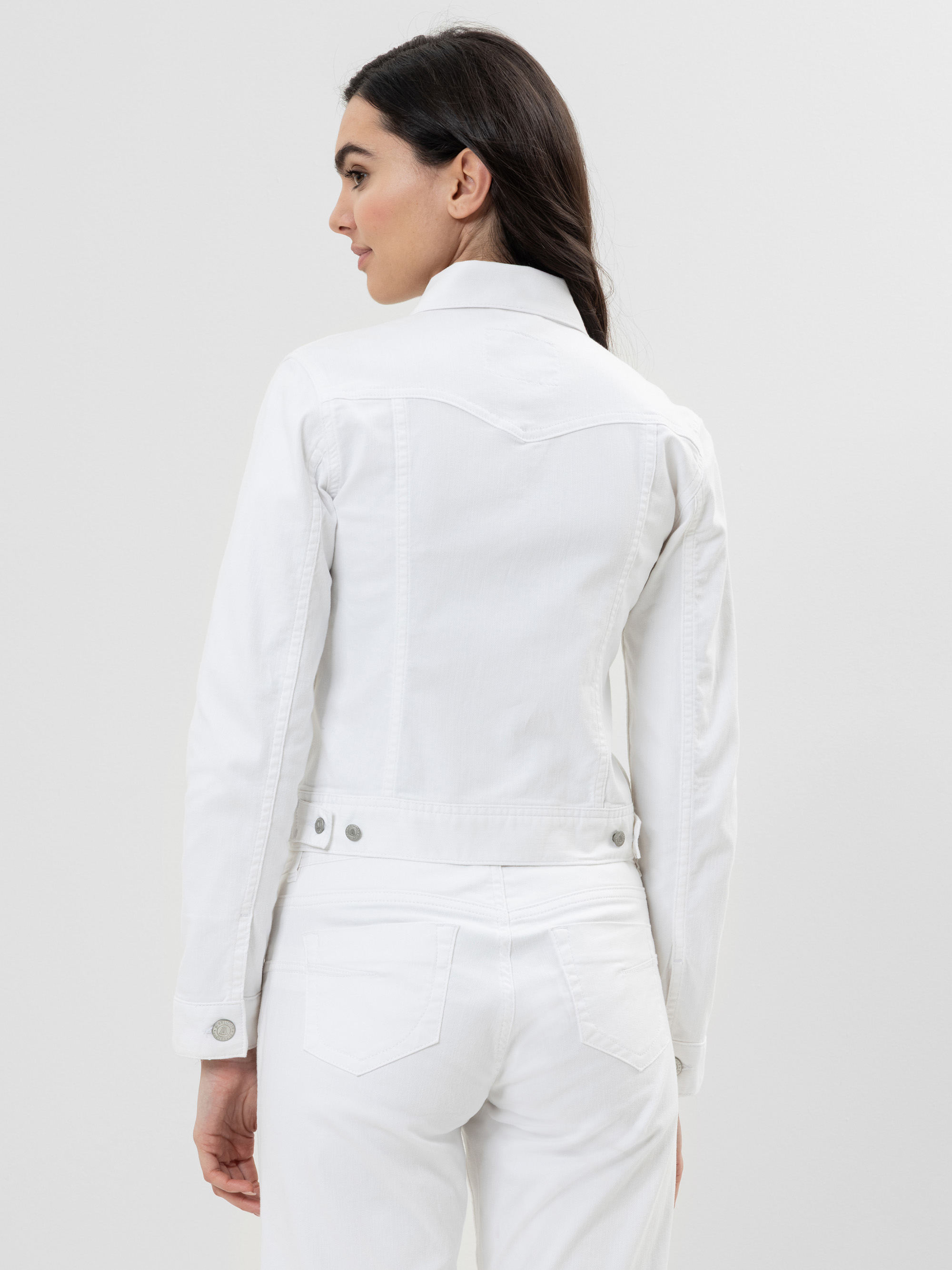 Eine Frau mit langen dunklen Haaren wird von hinten gezeigt. Sie trägt eine weiße Jeansjacke und eine weiße Hose vor einem schlichten hellen Hintergrund.