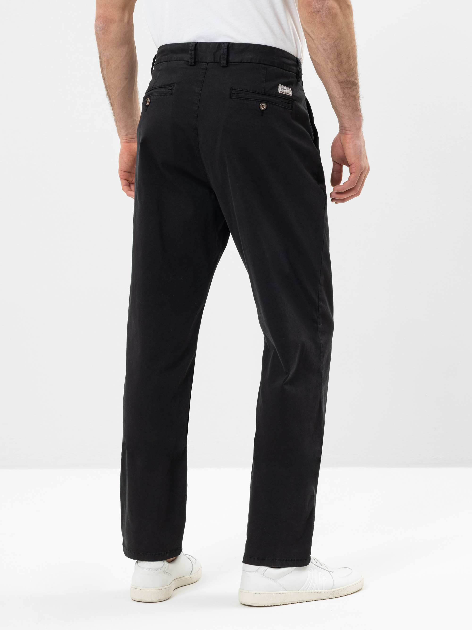 Ein Mann, der eine schwarze Hose, ein weißes T-Shirt und weiße Turnschuhe trägt, steht mit dem Gesicht nach hinten und zeigt die Rückenansicht der Hose in einer minimalen, hellen Studioumgebung.