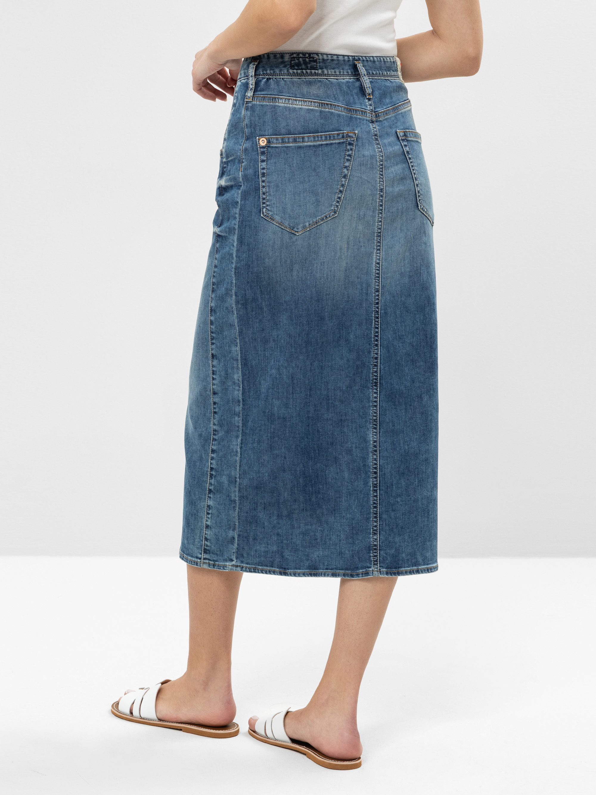 Eine Frau, die einen hellblauen Jeans-Midirock mit Gesäßtasche, ein weißes Oberteil und weiße Slide-Sandalen trägt, steht vor einem schlichten weißen Hintergrund.