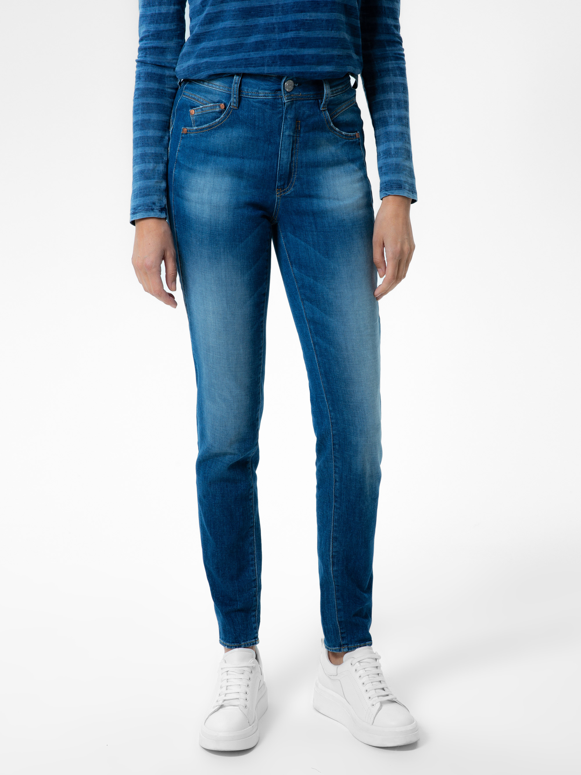 Person, die blaue Jeans, ein blaues langärmeliges gestreiftes Oberteil und weiße Turnschuhe trägt und vor einem schlichten weißen Hintergrund steht. Das Bild zeigt sie von den Schultern bis zu den Füßen.