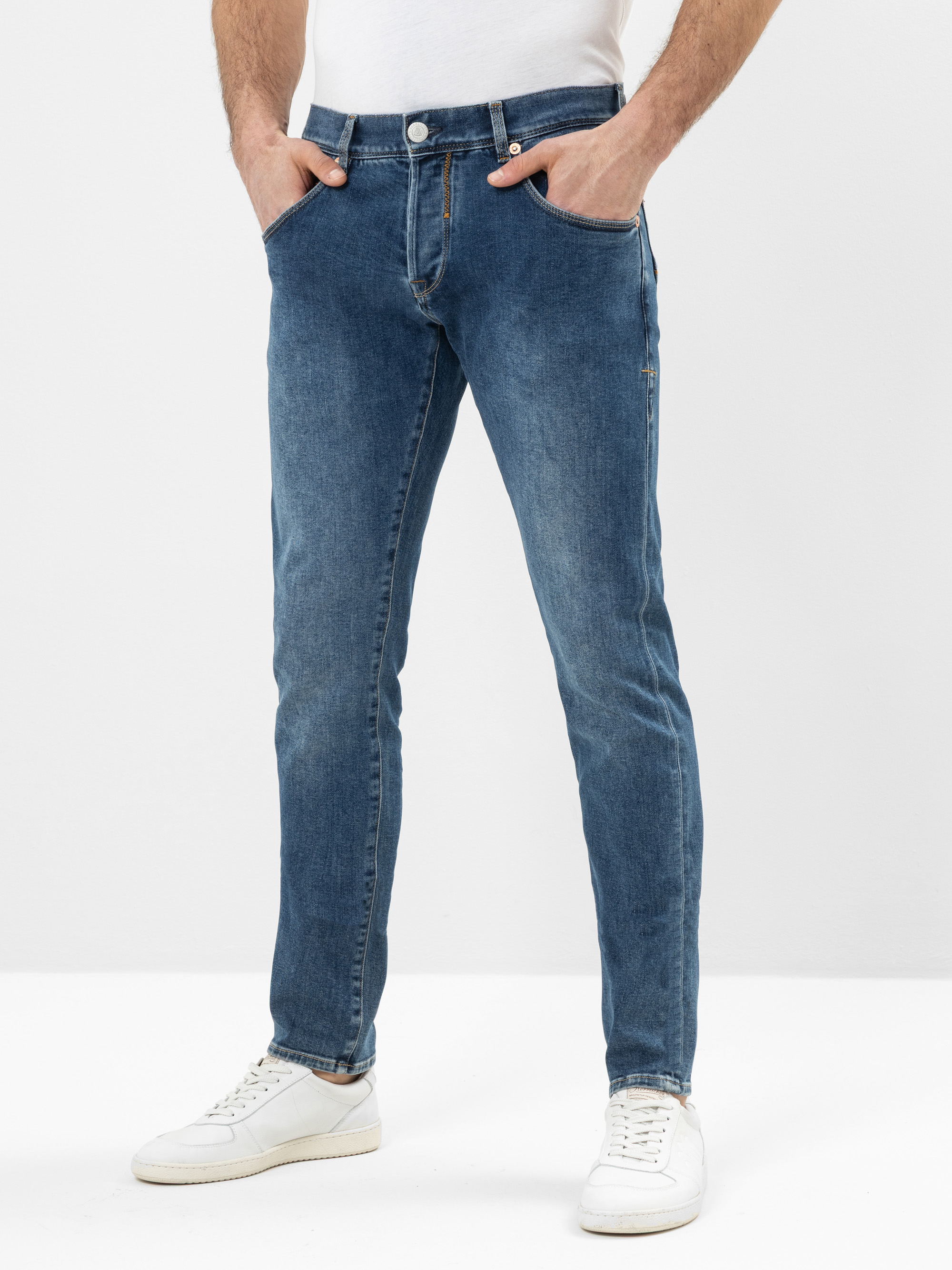 Eine Person, die mittelblaue Slim-Fit-Jeans und weiße Turnschuhe trägt, steht mit den Händen in den Taschen vor einem schlichten weißen Hintergrund.