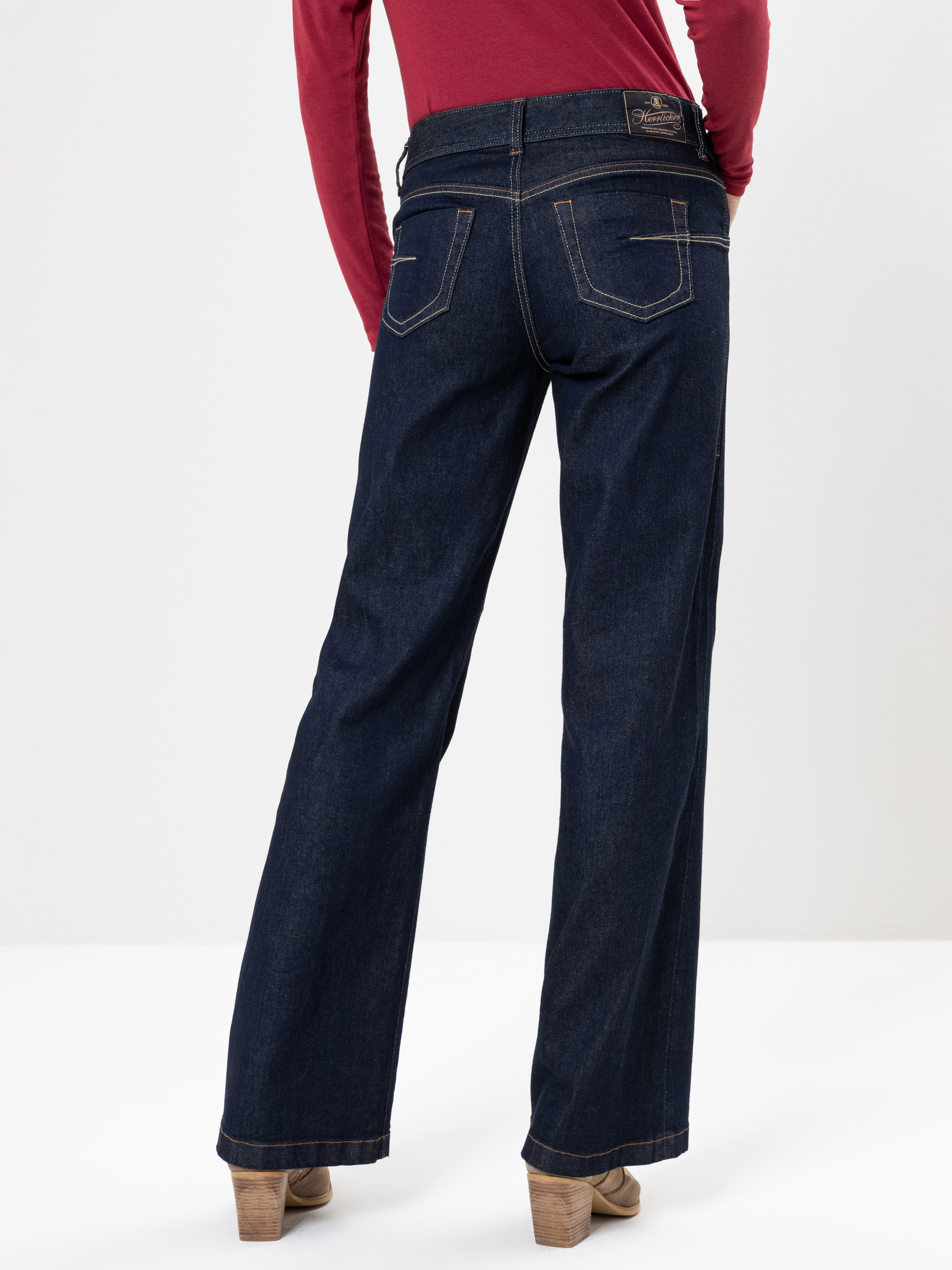 Eine Person, die dunkelblaue Jeans mit weitem Bein, ein langärmeliges rotes Oberteil und braune Schuhe mit Absätzen trägt, steht mit dem Rücken zur Kamera vor einem schlichten weißen Hintergrund.