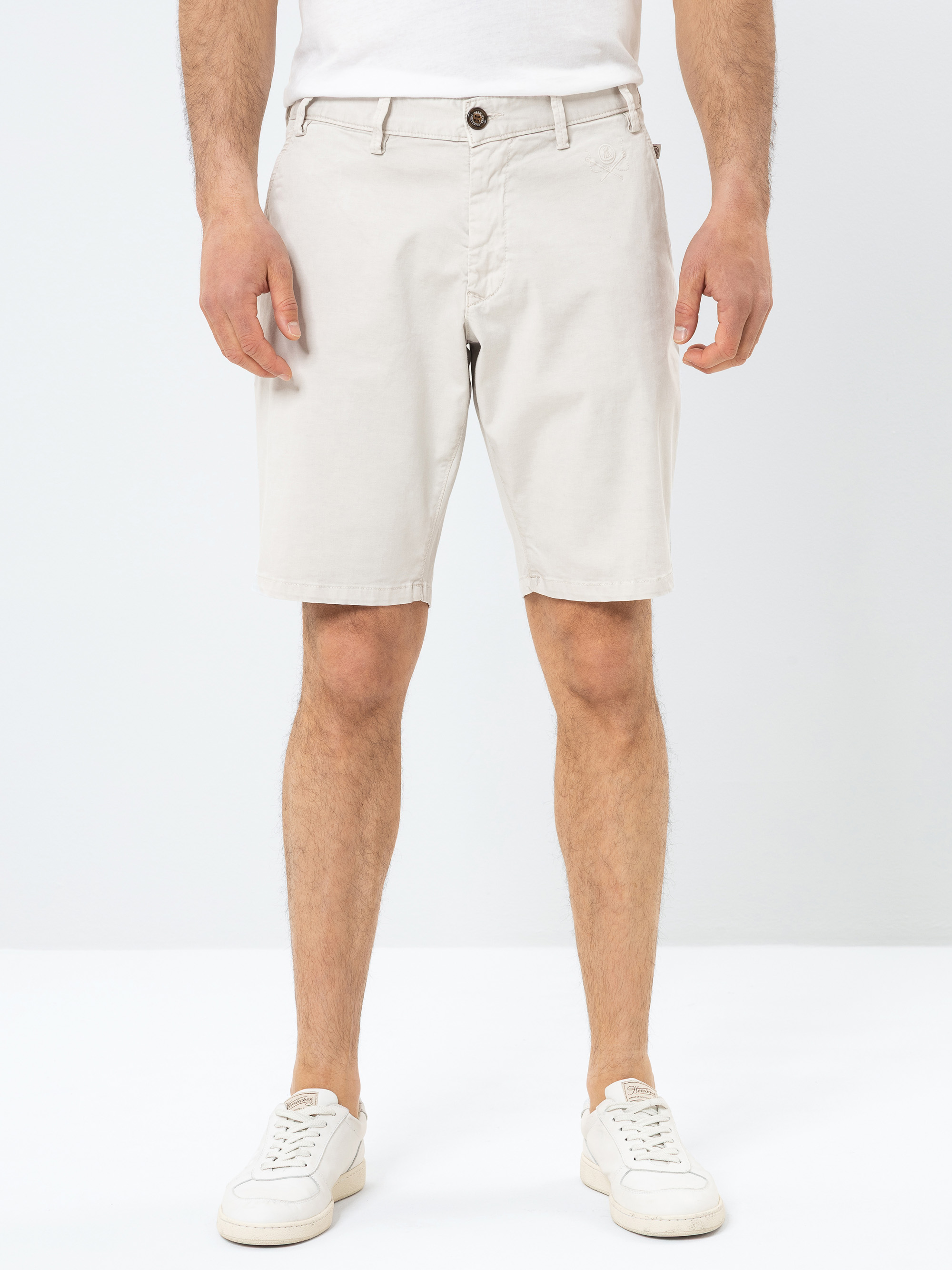 Ein Mann, der ein weißes T-Shirt, hellbeige knielange Shorts und weiße Turnschuhe trägt, steht vor einem schlichten weißen Hintergrund. Nur sein unterer Oberkörper und seine Beine sind sichtbar.