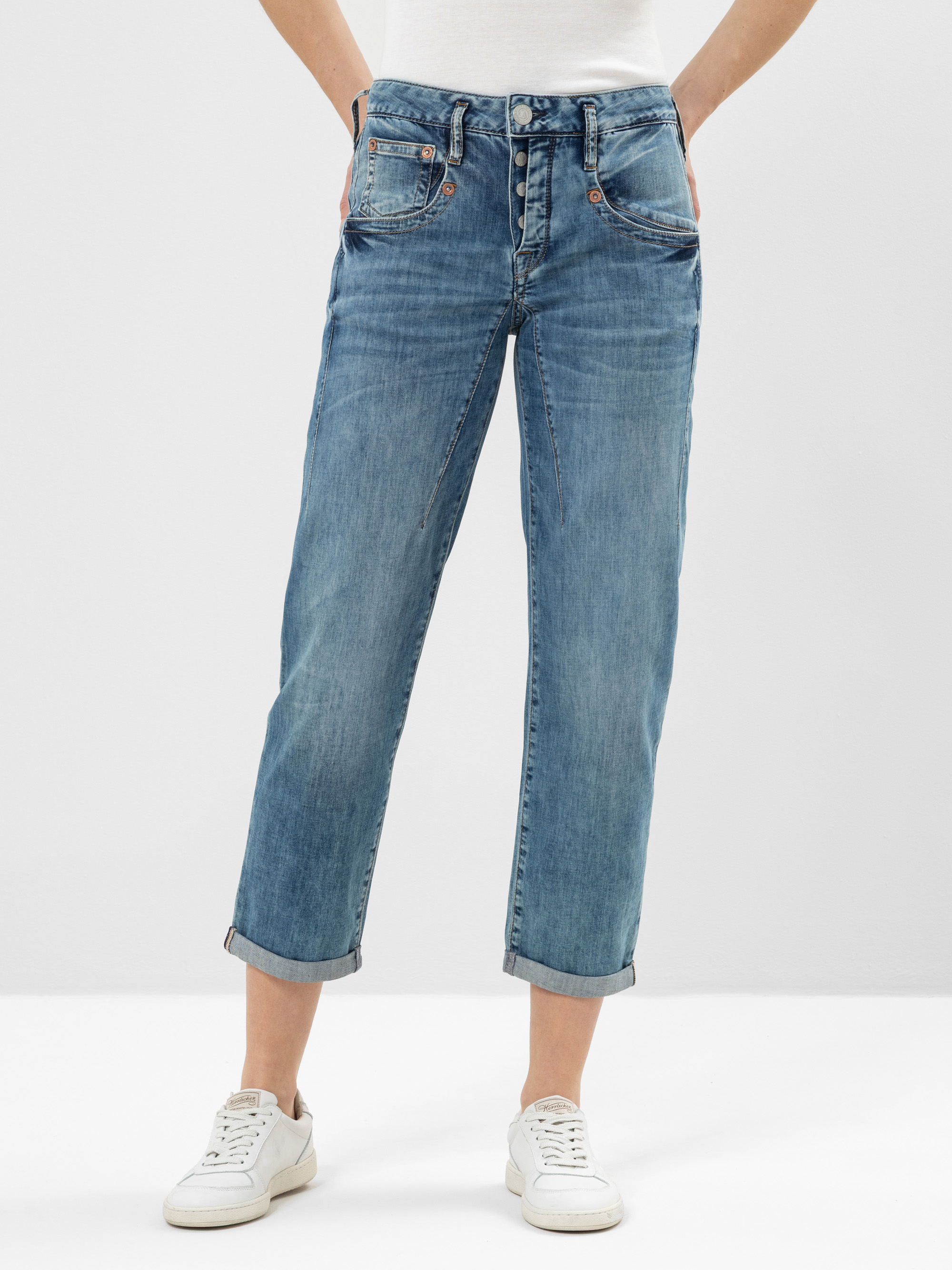 Eine Person, die hellblaue, mit Bündchen versehene, locker sitzende Jeans und weiße Turnschuhe trägt, steht mit den Händen in den Taschen vor einem schlichten Hintergrund.