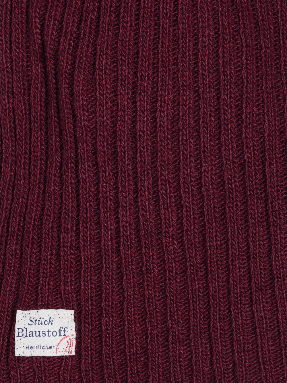 Nahaufnahme eines kastanienbraunen Rippenstrickstoffs mit einem kleinen weißen Etikett in der unteren Ecke, das deutschen Text und eine rote Markierung enthält.