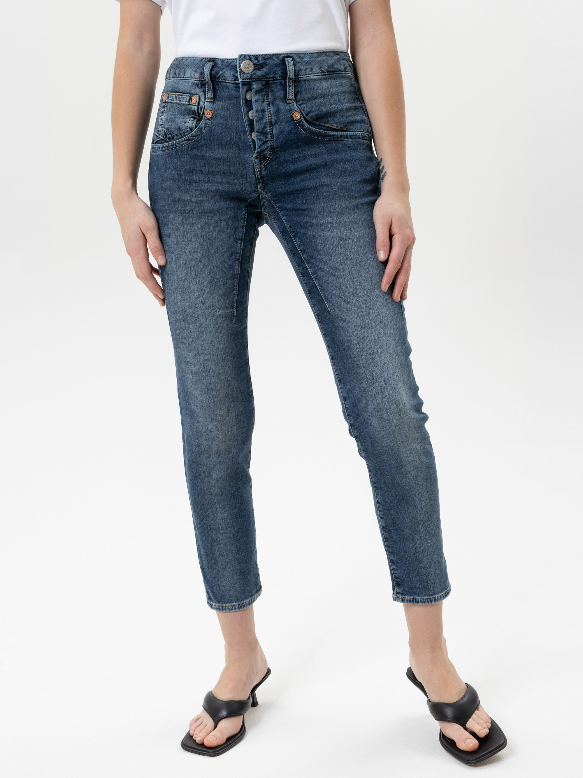 Eine Person, die ein weißes T-Shirt, hochgeschnittene blaue Röhrenjeans und schwarze hochhackige Sandalen trägt, steht vor einem schlichten weißen Hintergrund. Das Bild ist von den Schultern abwärts beschnitten.