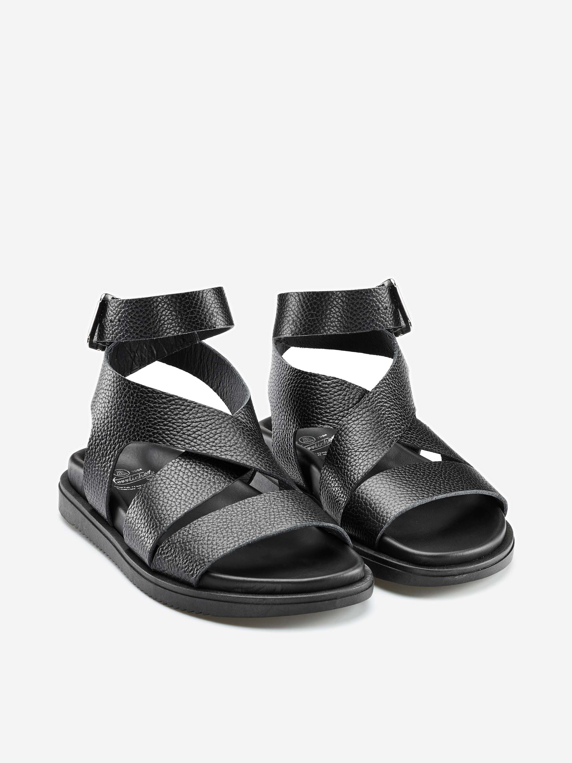Ein Paar schwarzer strukturierter Ledersandalen mit mehreren überkreuzten Riemen und Knöchelriemen mit Klettverschluss auf einem schlichten hellgrauen Hintergrund.